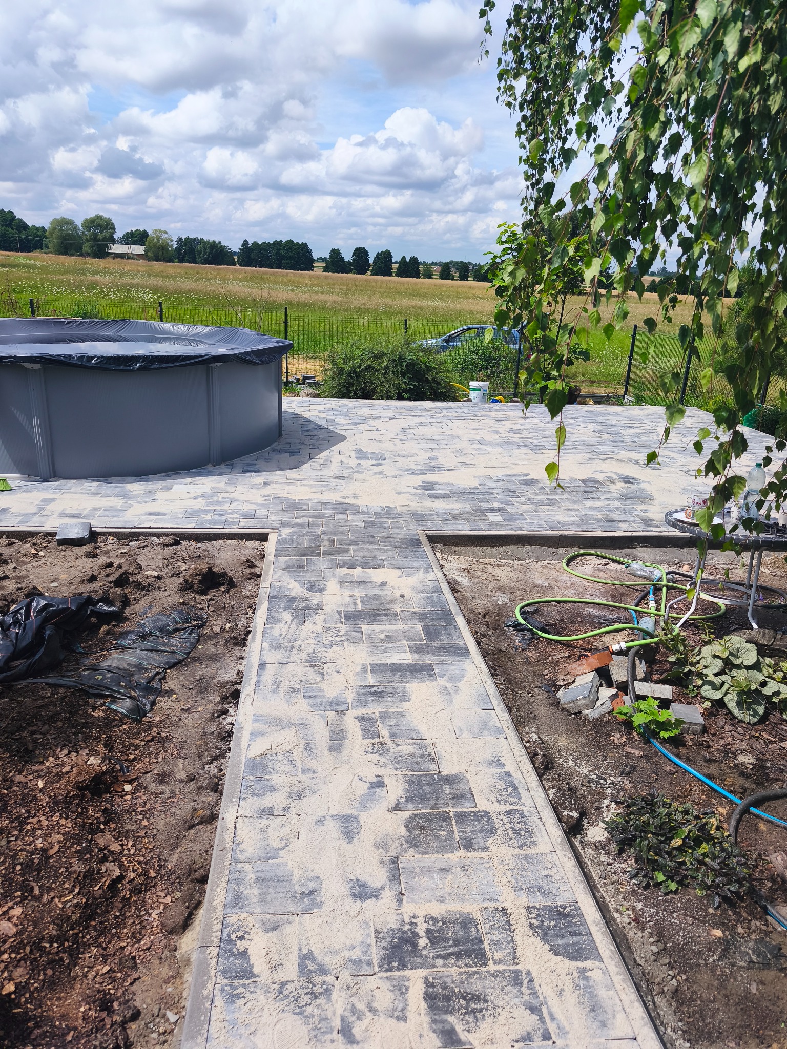 Układanie kostki brukowej na zewnątrz, widok na basen i otwartą przestrzeń z zielenią, w tle drzewa. Prace w toku, widoczne narzędzia i materiały budowlane.