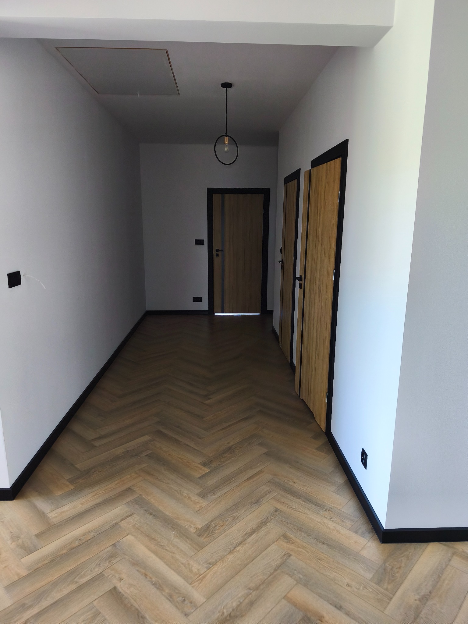 Korytarz po remoncie: parkiet w jodełkę, białe ściany z czarnymi listwami przypodłogowymi i ościeżnicami, nowoczesna lampa wisząca, dwoje drzwi.