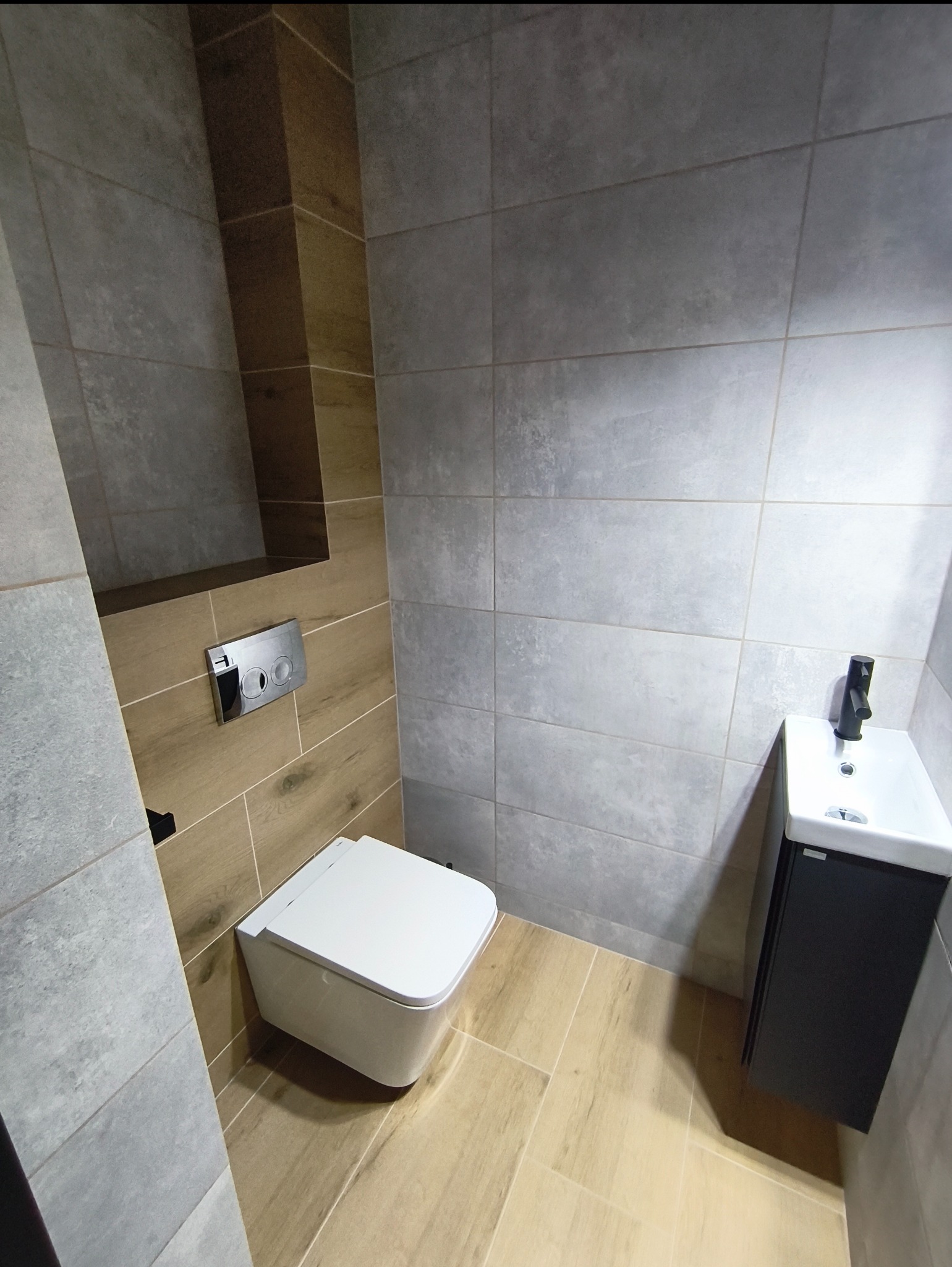 Nowoczesna łazienka z podwieszaną toaletą i umywalką, wykończona szarymi i drewnopodobnymi płytkami. Minimalistyczny design, armatura w kolorze czarnym.