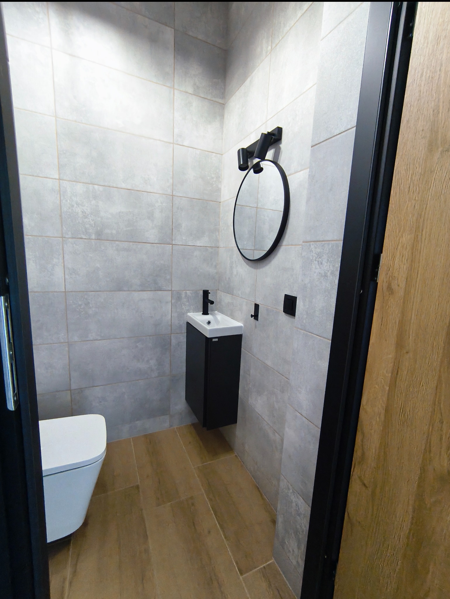 Nowoczesna łazienka w szarościach z drewnopodobną podłogą. Umywalka z czarną szafką, okrągłe lustro i wisząca toaleta. Minimalistyczny design.