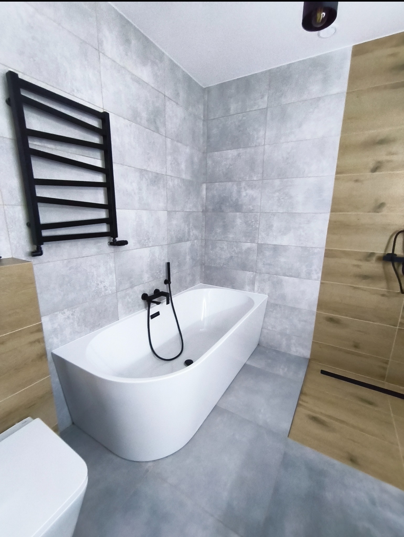 Nowoczesna łazienka z wolnostojącą wanną, grafitową armaturą i grzejnikiem drabinkowym na tle szarych płytek. Uwagę zwraca drewnopodobna okładzina.