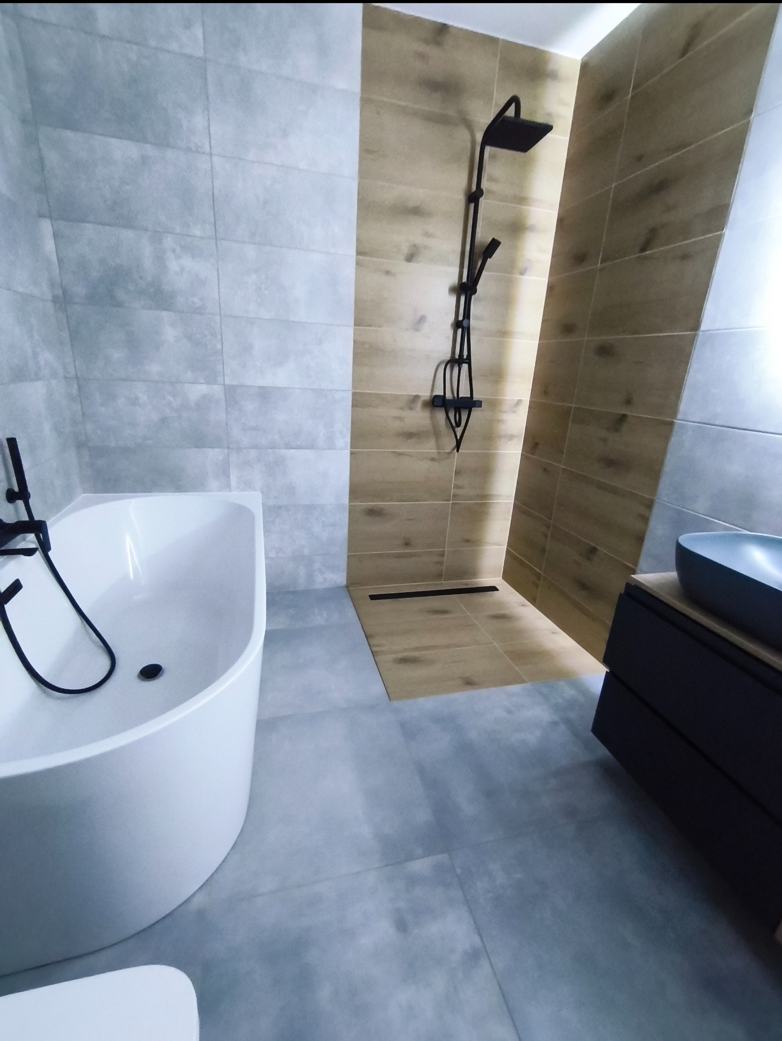 Nowoczesna łazienka z wanną, kabiną prysznicową typu walk-in i szarą płytką podłogową. Czarna armatura i drewnopodobne panele ścienne dodają elegancji.