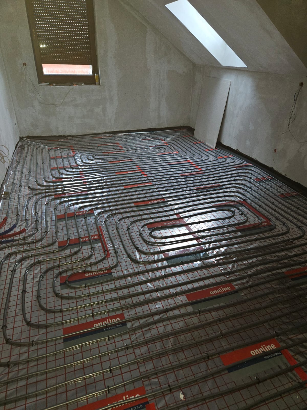 Ułożona instalacja ogrzewania podłogowego w surowym wnętrzu poddasza, widoczne rury grzewcze na folii z siatką, okno dachowe i okno w ścianie. Etap przed wylewką.