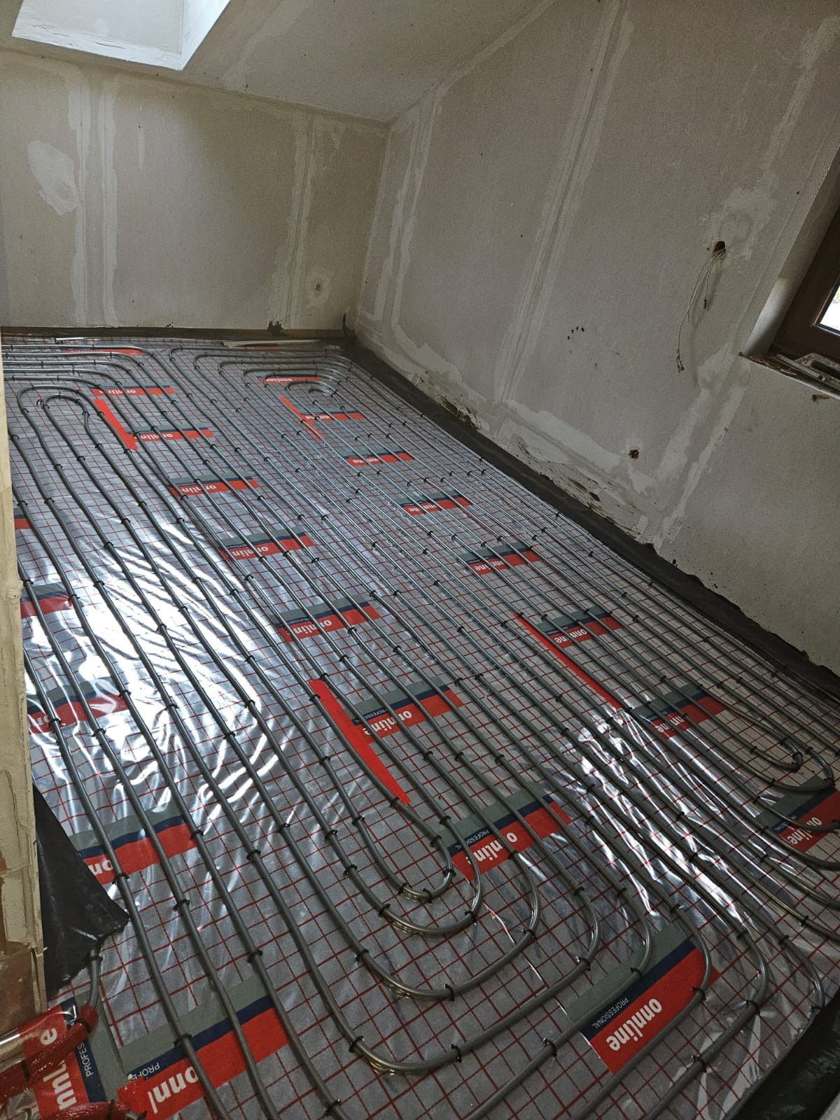 Instalacja ogrzewania podłogowego z rurkami na folii w surowym wnętrzu poddasza. Widoczne elementy montażowe i przygotowane podłoże.