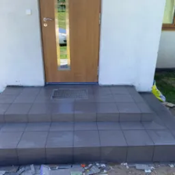 Wykończone płytkami schody wejściowe z kratką odpływową przed drzwiami wejściowymi. Widoczne resztki materiałów budowlanych.