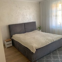 Usługi Budowlane Roman Borsuk - Wykończona sypialnia: szara tapicerowana rama łóżka z kremowym pledem, drewniana podłoga i dekoracyjna ściana w jasnych odcieniach. Okno z firanami w tle.