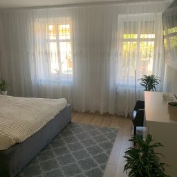 Usługi Budowlane Roman Borsuk - Jasna sypialnia z łóżkiem, szarym dywanem i białymi, zwiewnymi zasłonami w oknach. Drewniana podłoga i zielone rośliny dodają przytulności wnętrzu.