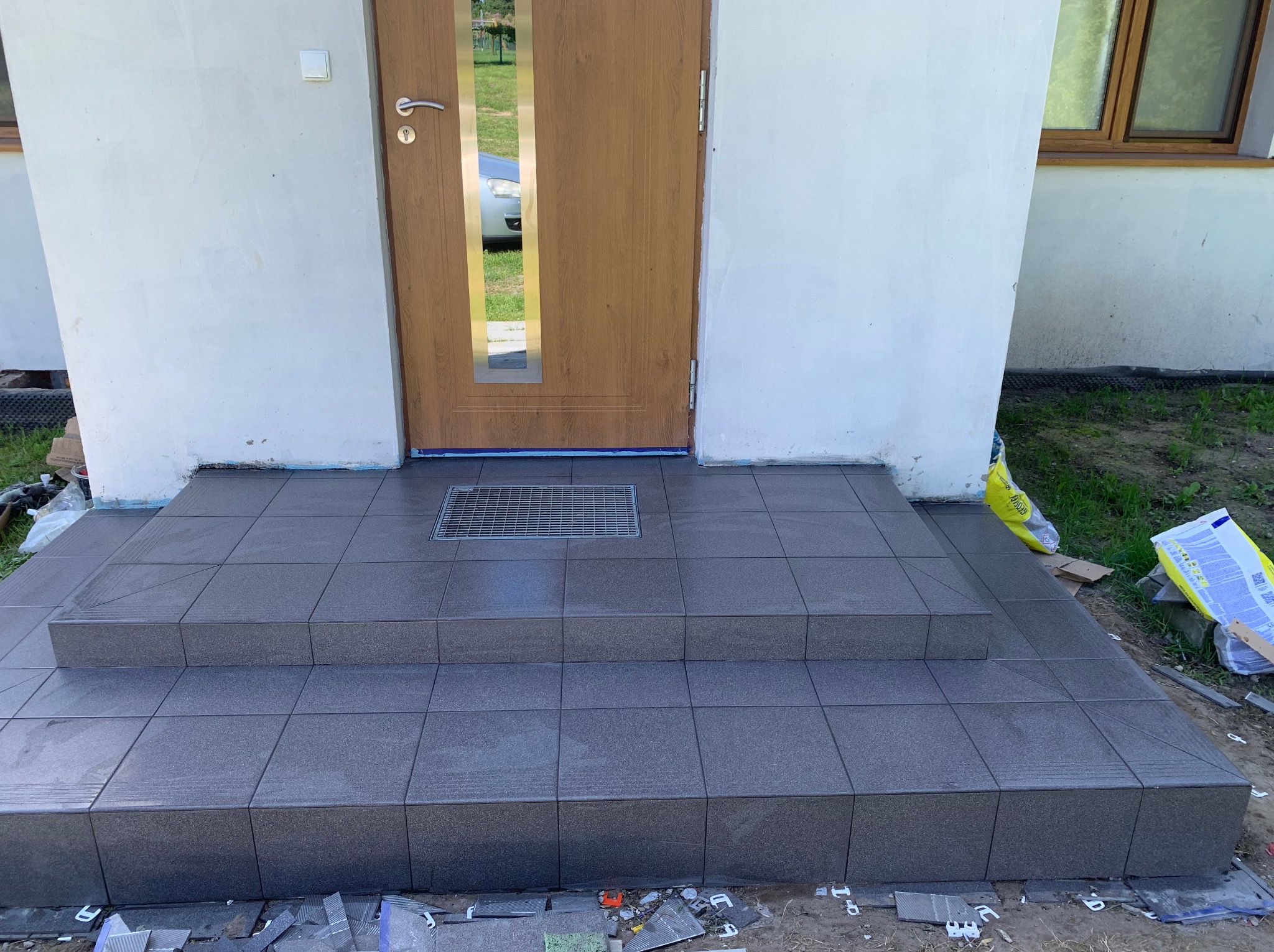 Wykończone płytkami schody wejściowe z kratką odpływową przed drzwiami wejściowymi. Widoczne resztki materiałów budowlanych.