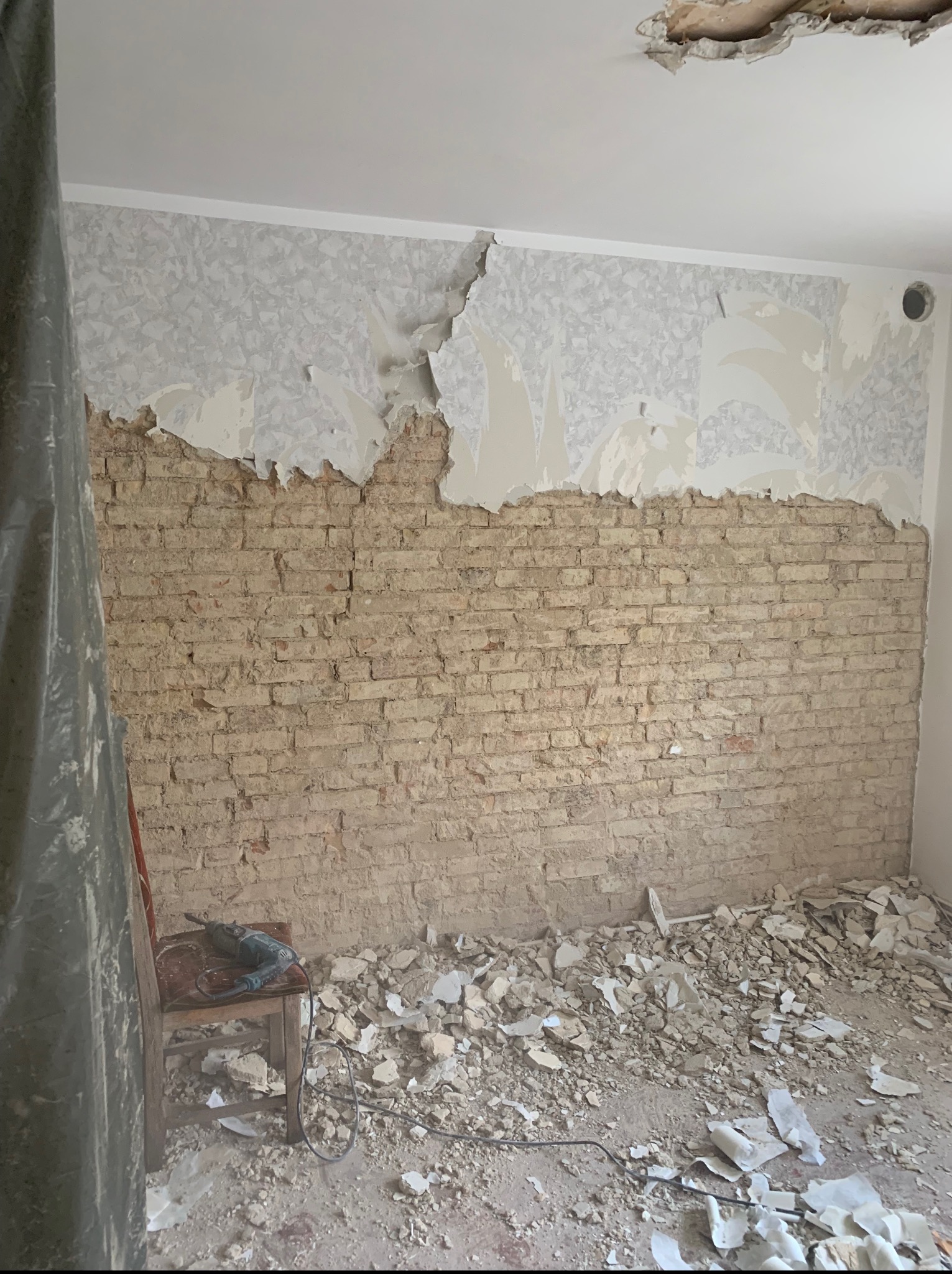 Odsłonięta ceglana ściana w trakcie skuwania tynku, widoczne resztki tapety, gruz na podłodze i wiertarka na krześle. Fragment zniszczonego sufitu.