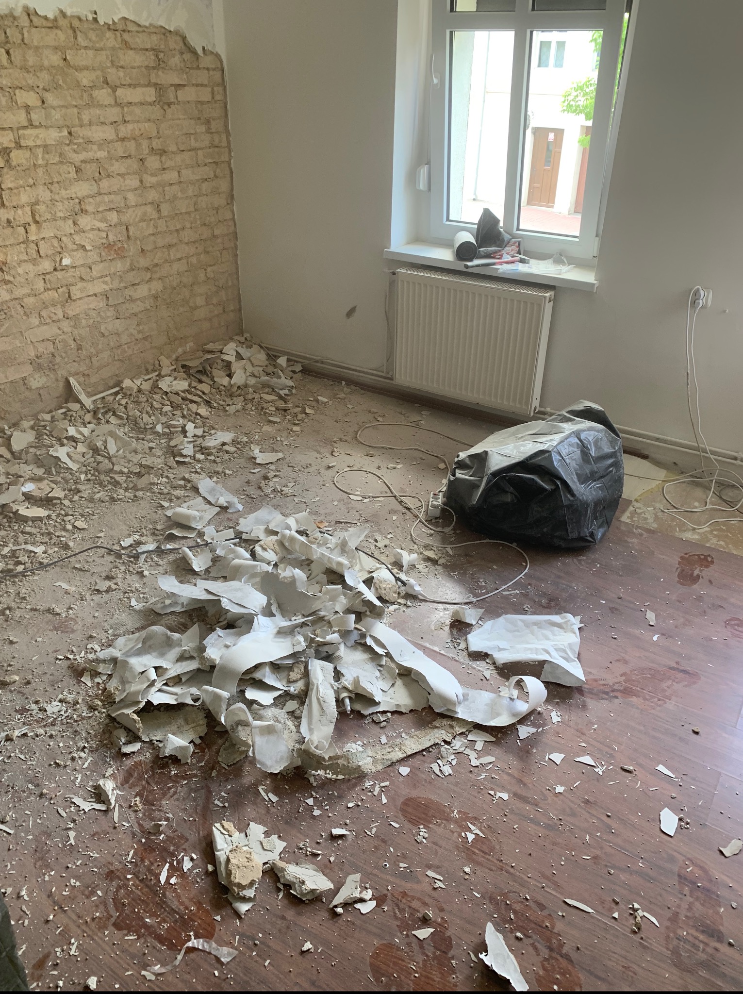 Pokój w trakcie remontu: odsłonięta cegła, gruz na podłodze, okno z widokiem na zewnątrz. Czarny worek na śmieci i resztki materiałów budowlanych.