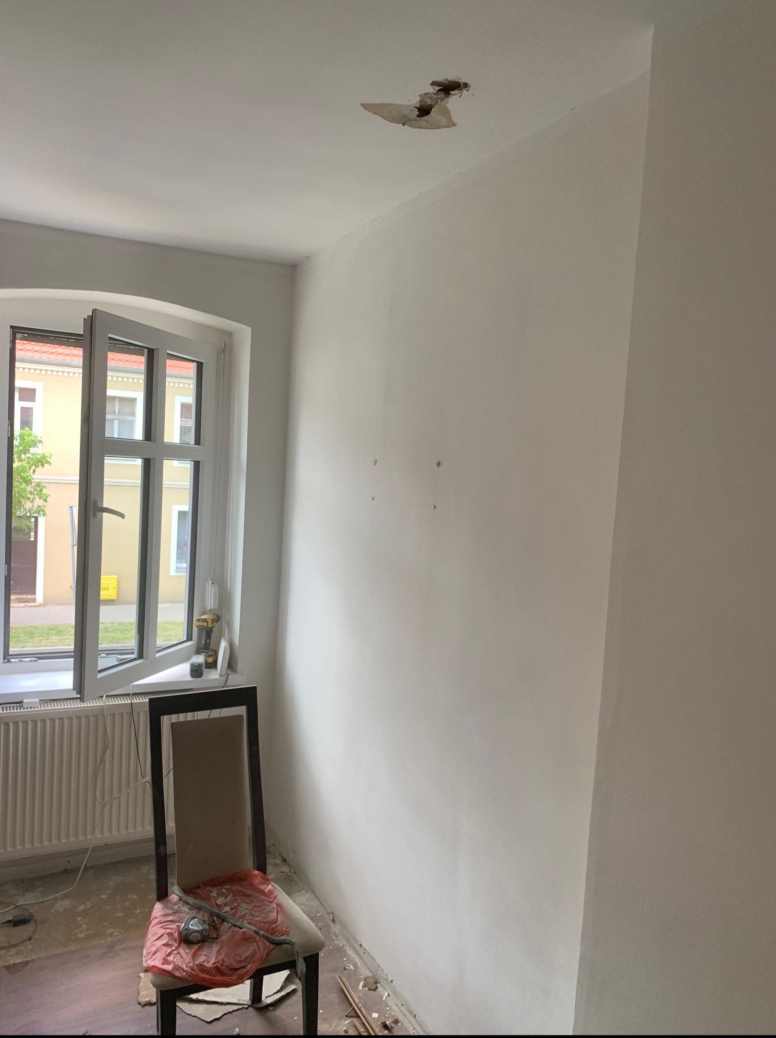 Wnętrze pokoju w trakcie odświeżania: gładkie, pomalowane ściany, okno z widokiem na ulicę, narzędzia na krześle. Fragment uszkodzonego sufitu w rogu.