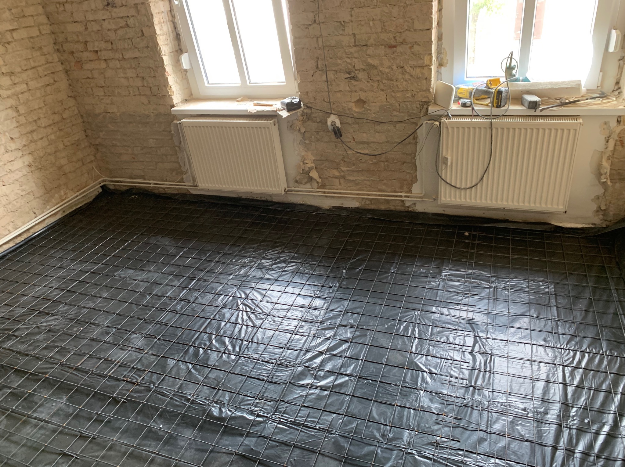 Przygotowanie podłoża: siatka zbrojeniowa na folii izolacyjnej w surowym wnętrzu z ceglanymi ścianami i grzejnikami, gotowe do wylewki.