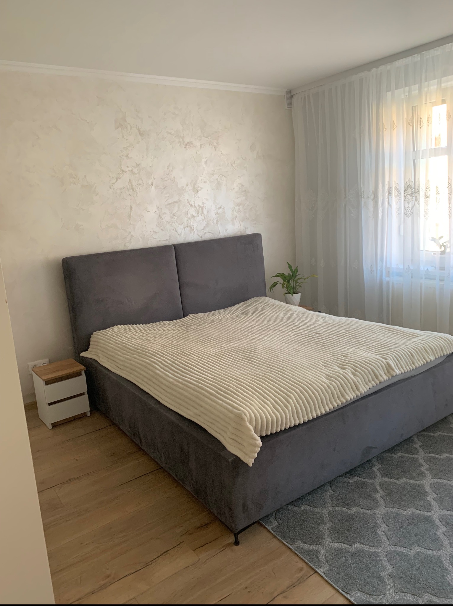 Wykończona sypialnia: szara tapicerowana rama łóżka z kremowym pledem, drewniana podłoga i dekoracyjna ściana w jasnych odcieniach. Okno z firanami w tle.