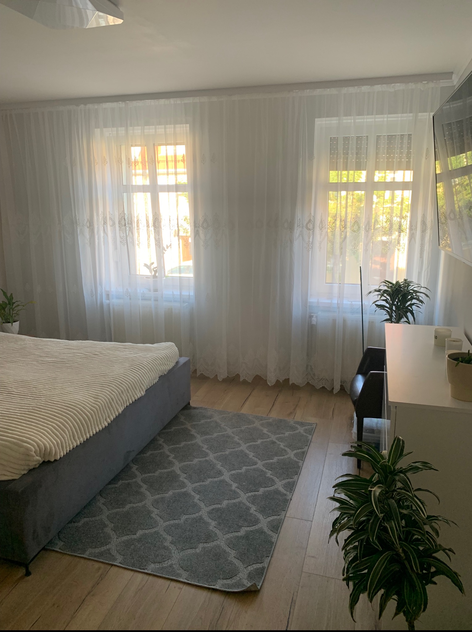 Jasna sypialnia z łóżkiem, szarym dywanem i białymi, zwiewnymi zasłonami w oknach. Drewniana podłoga i zielone rośliny dodają przytulności wnętrzu.