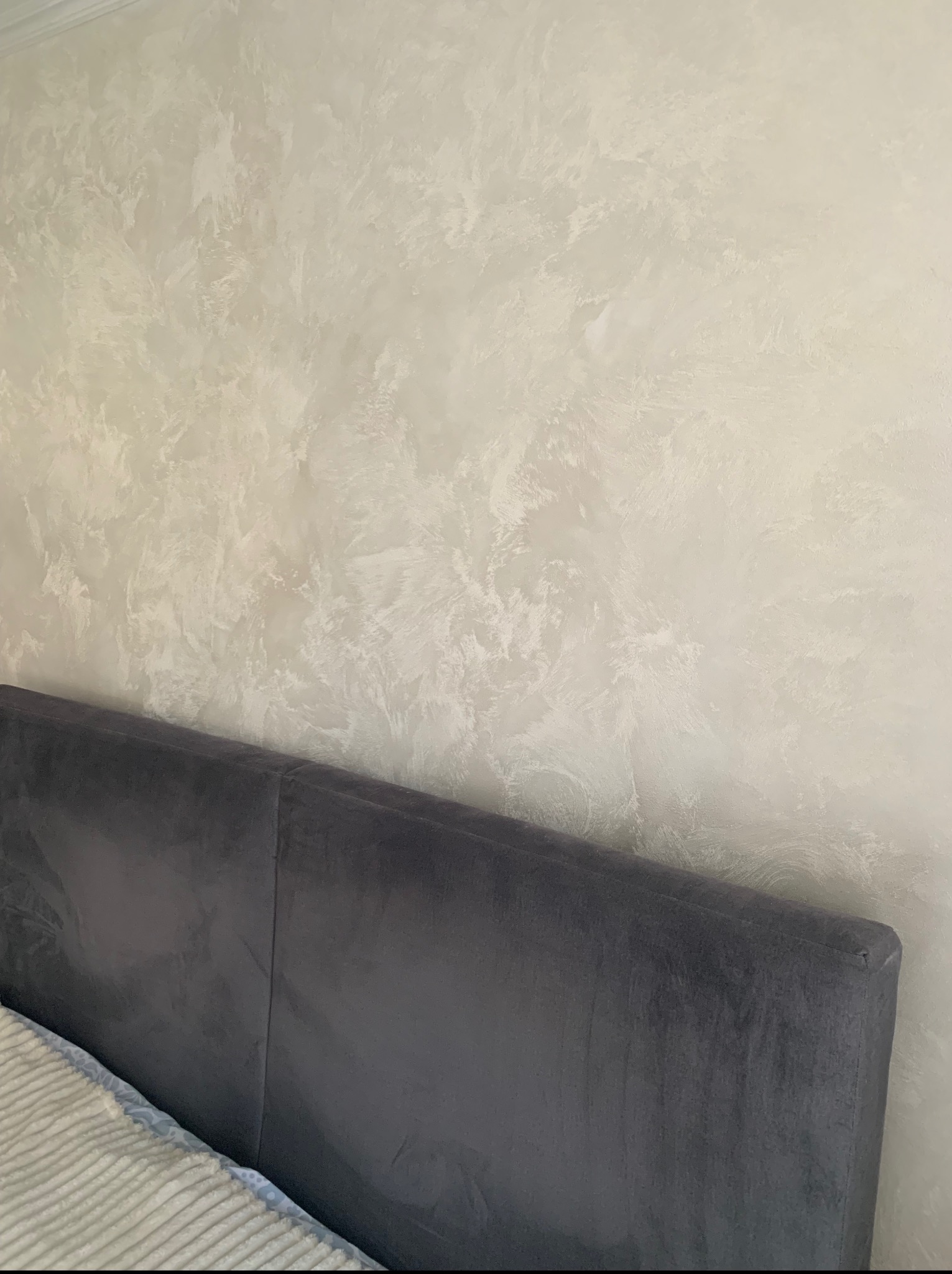 Dekoracyjna ściana w jasnych, kremowych odcieniach z efektem stiuku weneckiego, widoczny fragment szarego wezgłowia łóżka i pościeli w dolnej części kadru.