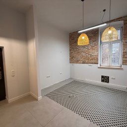 Mar Den - Wnętrze po remoncie: szaro-biała podłoga w szachownicę, ceglana ściana z oknem, nowoczesne lampy wiszące. Widoczne gniazdka elektryczne i rury pod oknem.