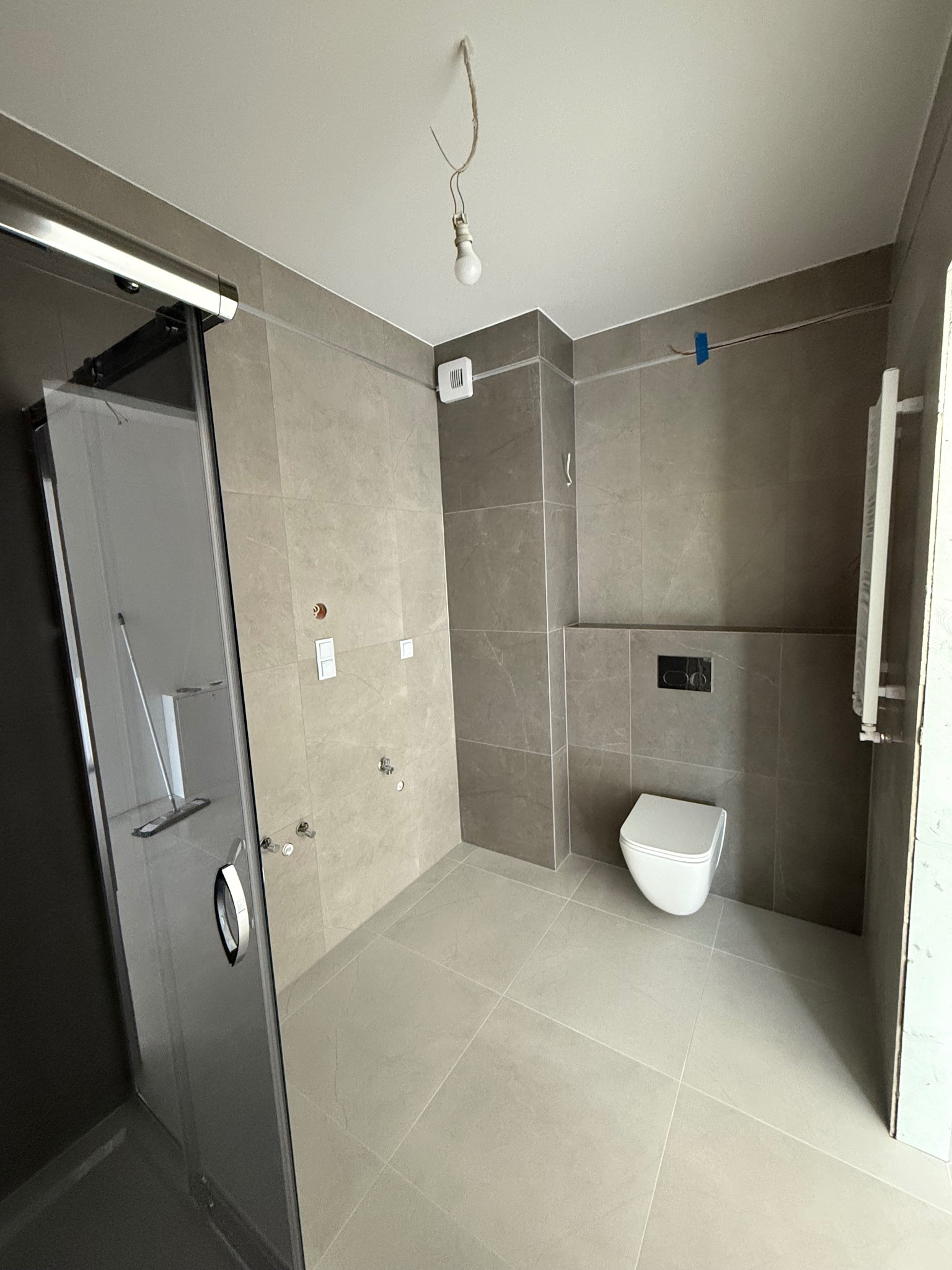 Wykończona łazienka z kabiną prysznicową, płytkami w odcieniach szarości, wiszącą toaletą i grzejnikiem. Widoczne instalacje elektryczne i oświetlenie tymczasowe.