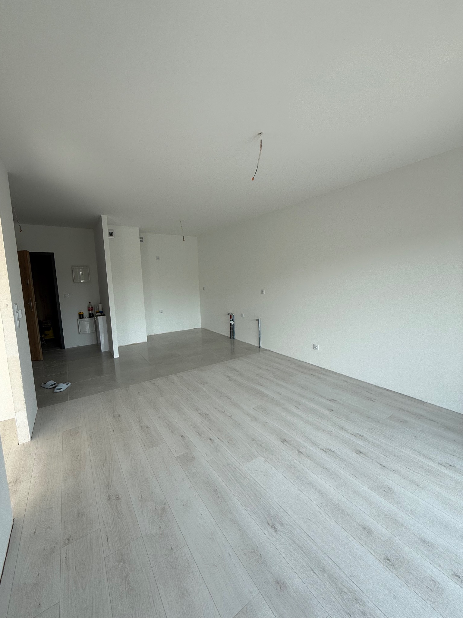 Jasne panele podłogowe w nowym wnętrzu. Widoczne gładkie, białe ściany i fragmenty instalacji. W tle płytki i wyposażenie łazienki. Minimalistyczne wykończenie.