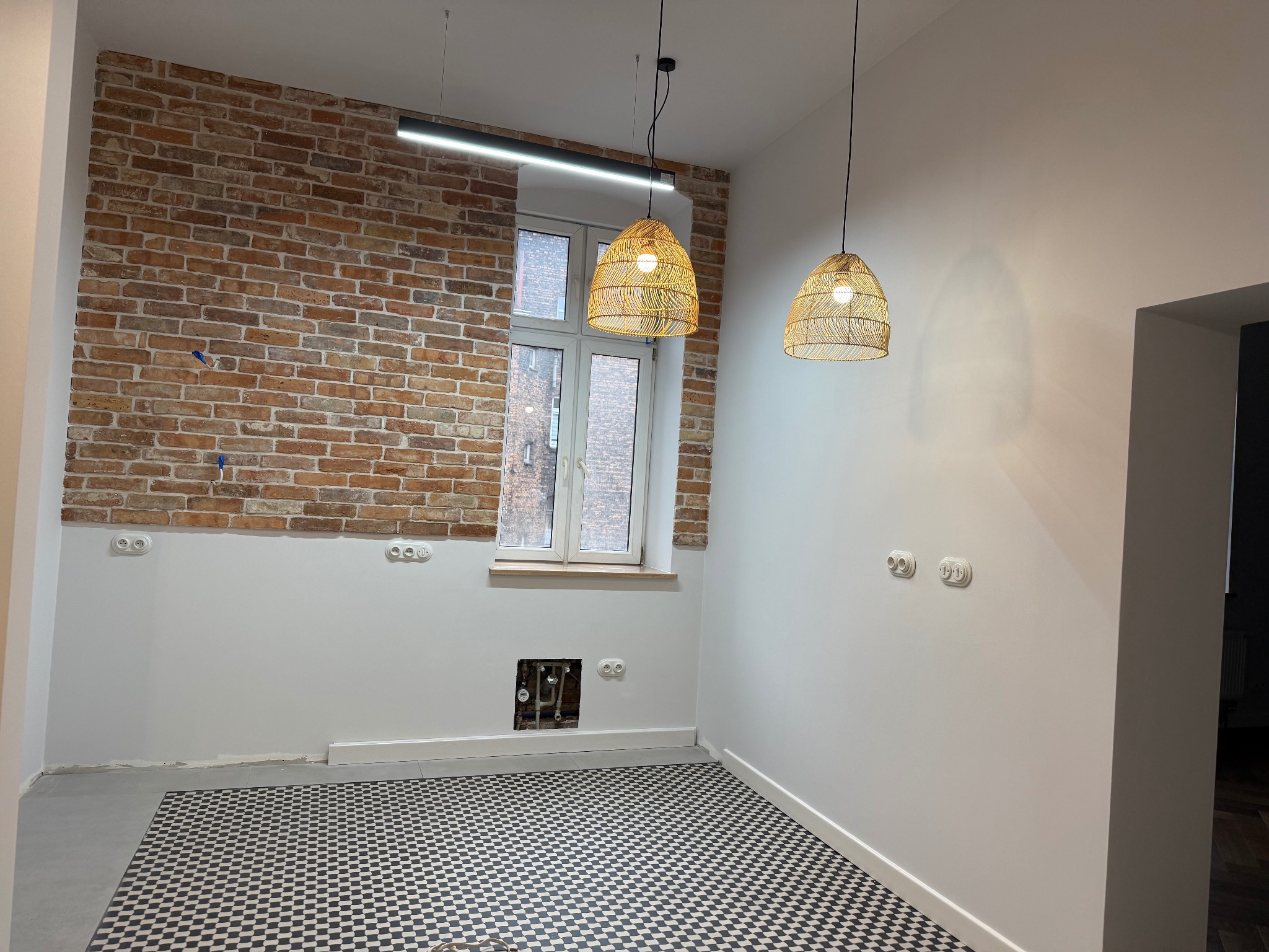 Wykończona kuchnia w stylu retro w Chorzowie: ceglana ściana, białe ściany, czarno-biała podłoga, okno i lampy wiszące. Widoczne gniazdka elektryczne i instalacja wodna.