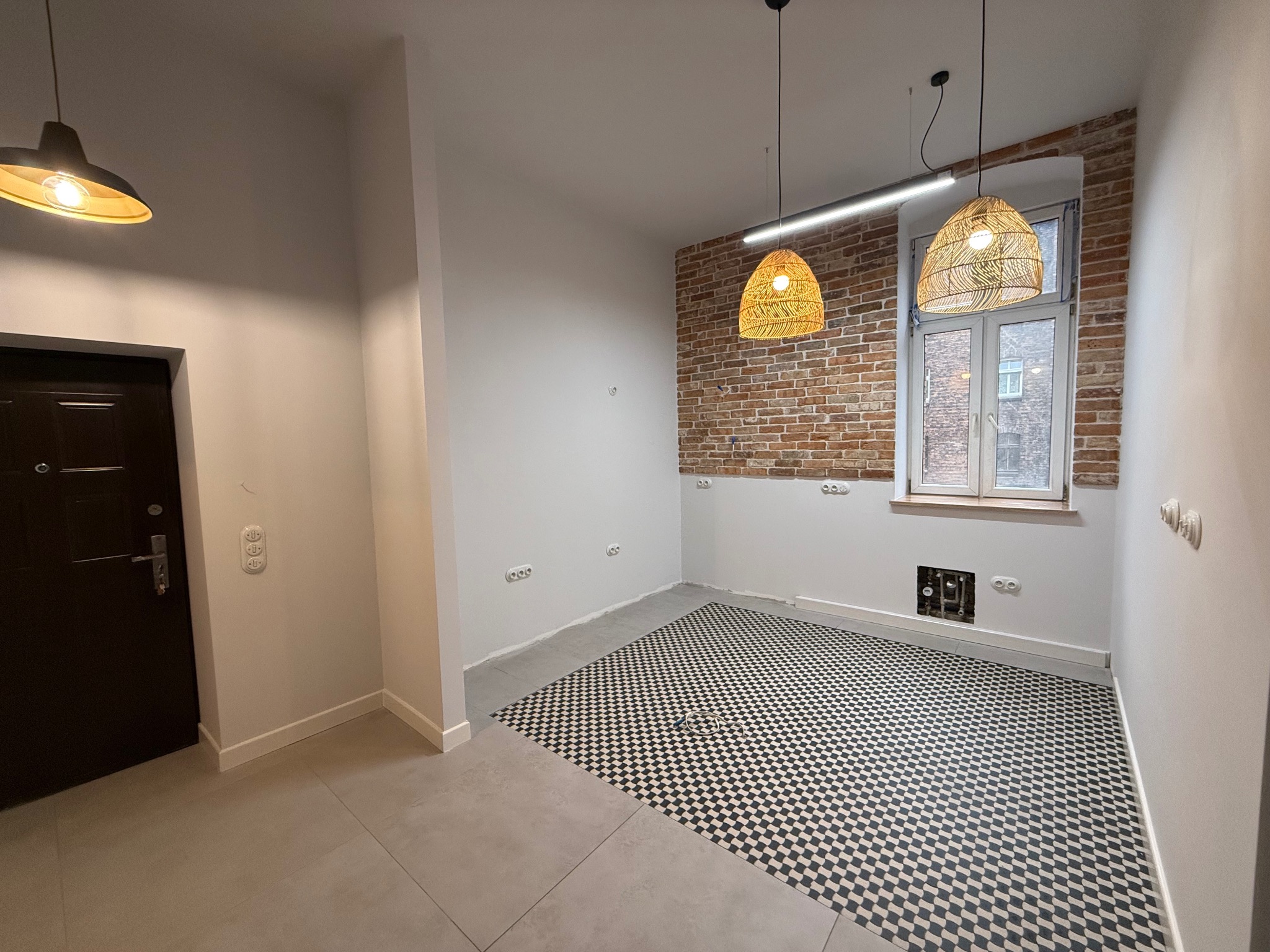 Wnętrze po remoncie: szaro-biała podłoga w szachownicę, ceglana ściana z oknem, nowoczesne lampy wiszące. Widoczne gniazdka elektryczne i rury pod oknem.