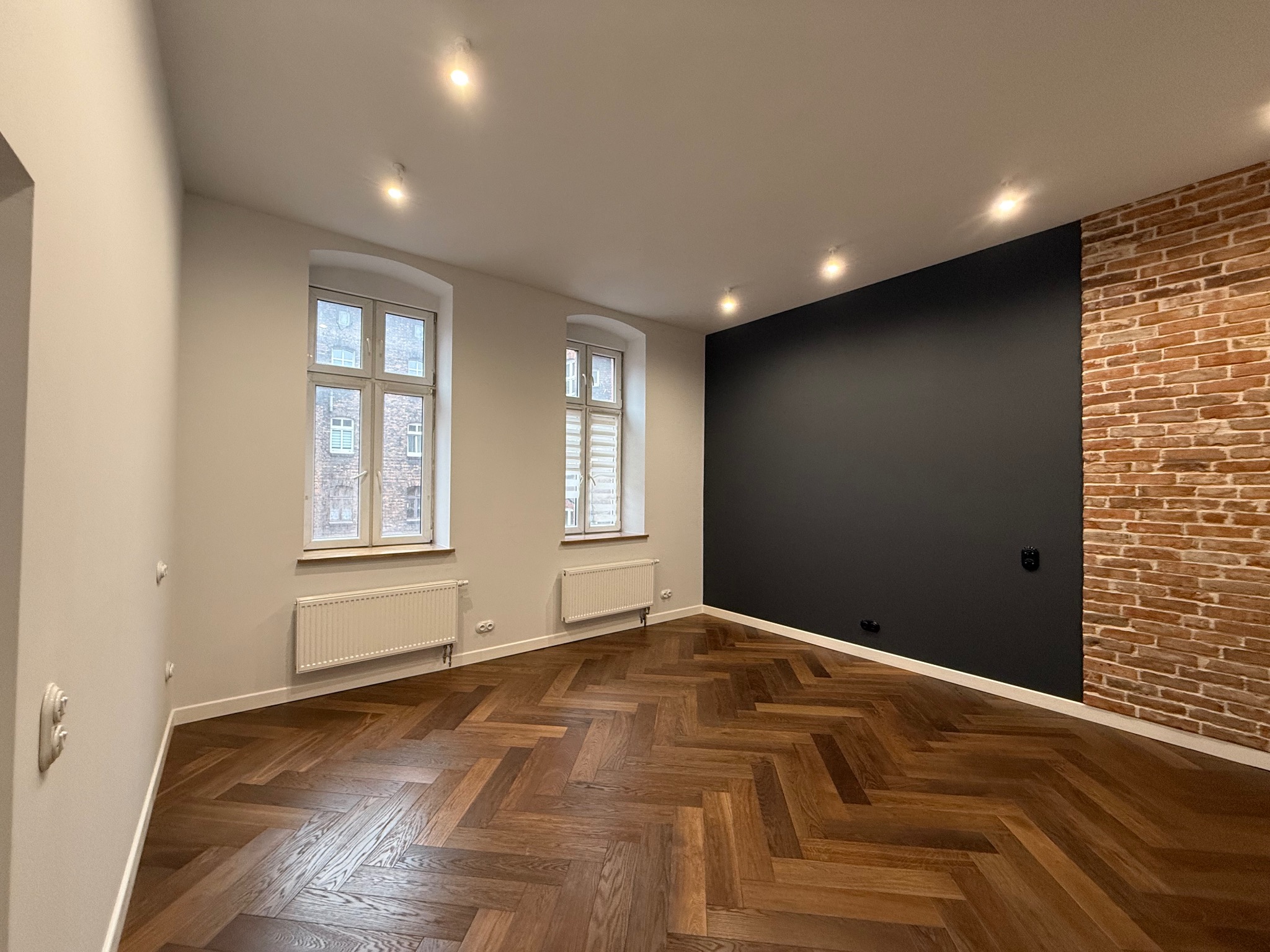 Wnętrze po remoncie: parkiet w jodełkę, ściana z cegły kontrastująca z czarną, dwa okna z grzejnikami. Nowoczesne oświetlenie punktowe.