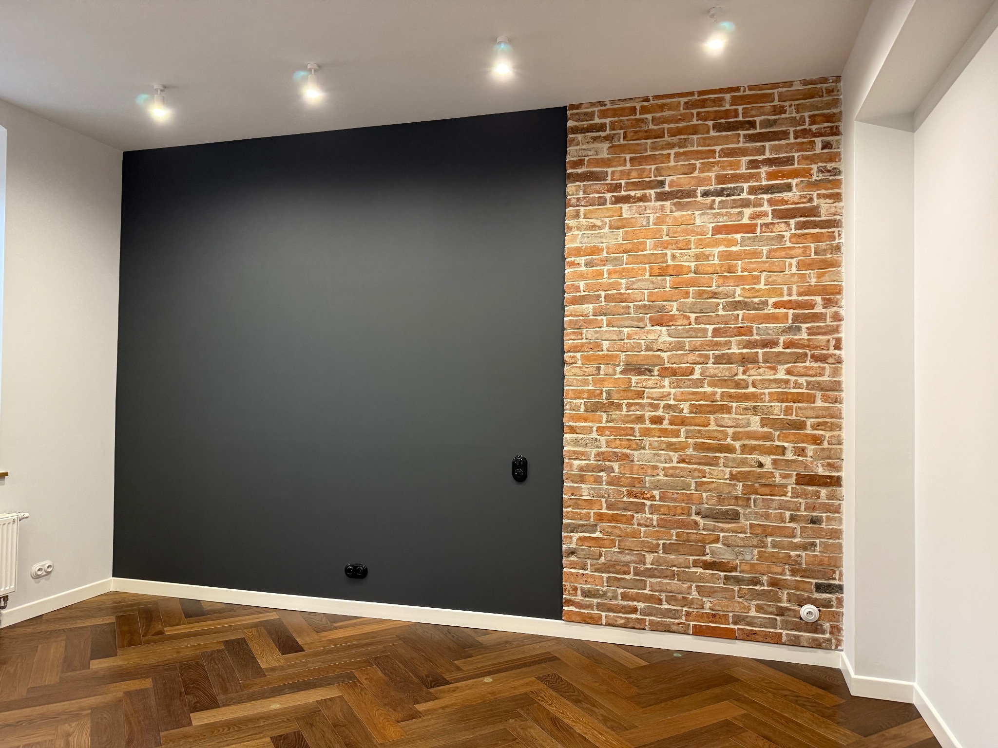 Wnętrze po remoncie: ciemnoszara ściana sąsiaduje z fragmentem ściany z cegły. Parkiet ułożony w jodełkę. Białe listwy przypodłogowe i sufit z oświetleniem punktowym.