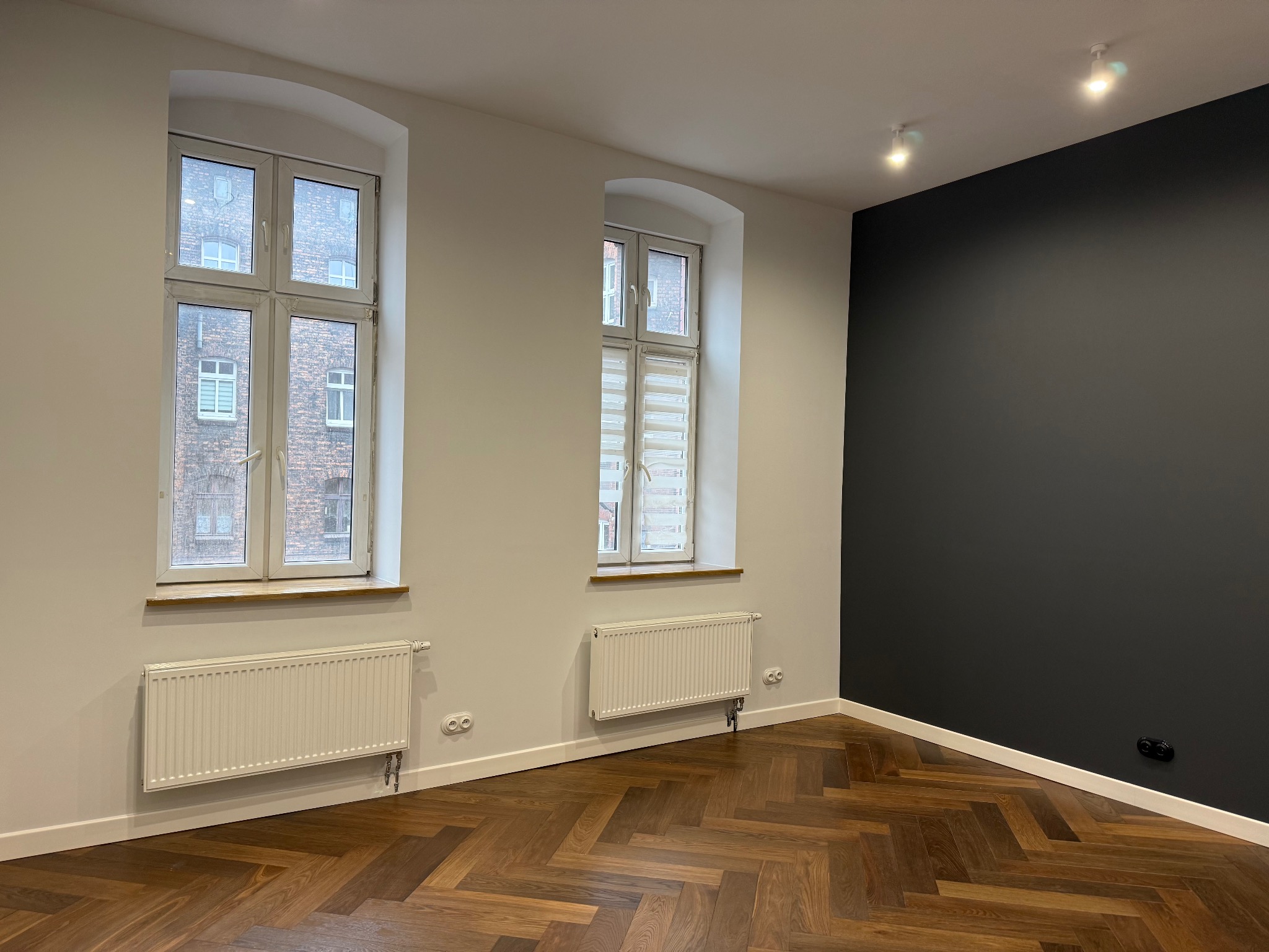 Wnętrze po remoncie: parkiet w jodełkę, białe ściany z dwoma oknami z widokiem na budynek z cegły, czarna ściana akcentująca, białe grzejniki.