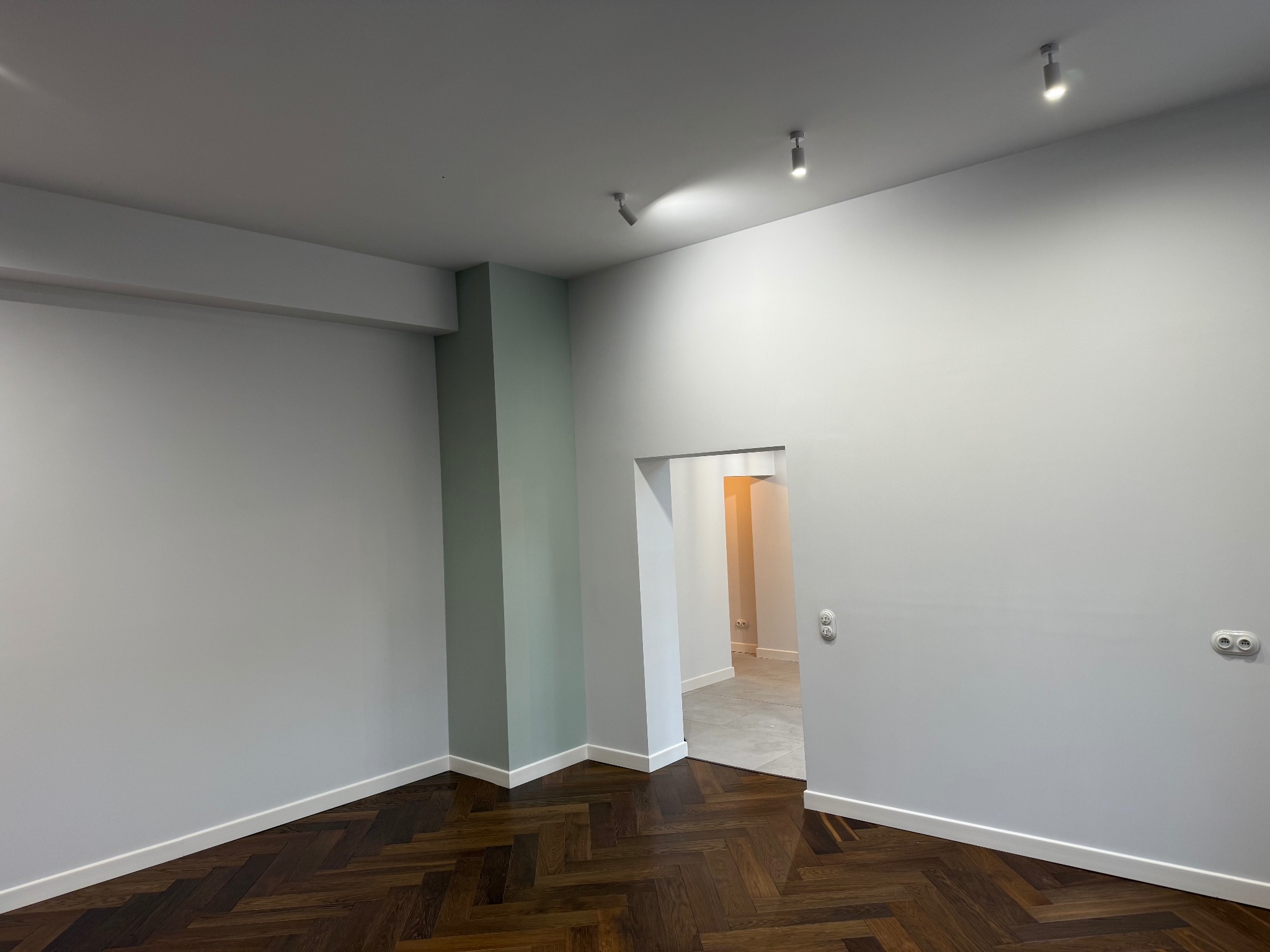 Pomalowane na szaro wnętrze z parkietem w jodełkę. Widoczne białe listwy przypodłogowe i włączniki, oraz oświetlenie punktowe. W tle widok na inne pomieszczenie.