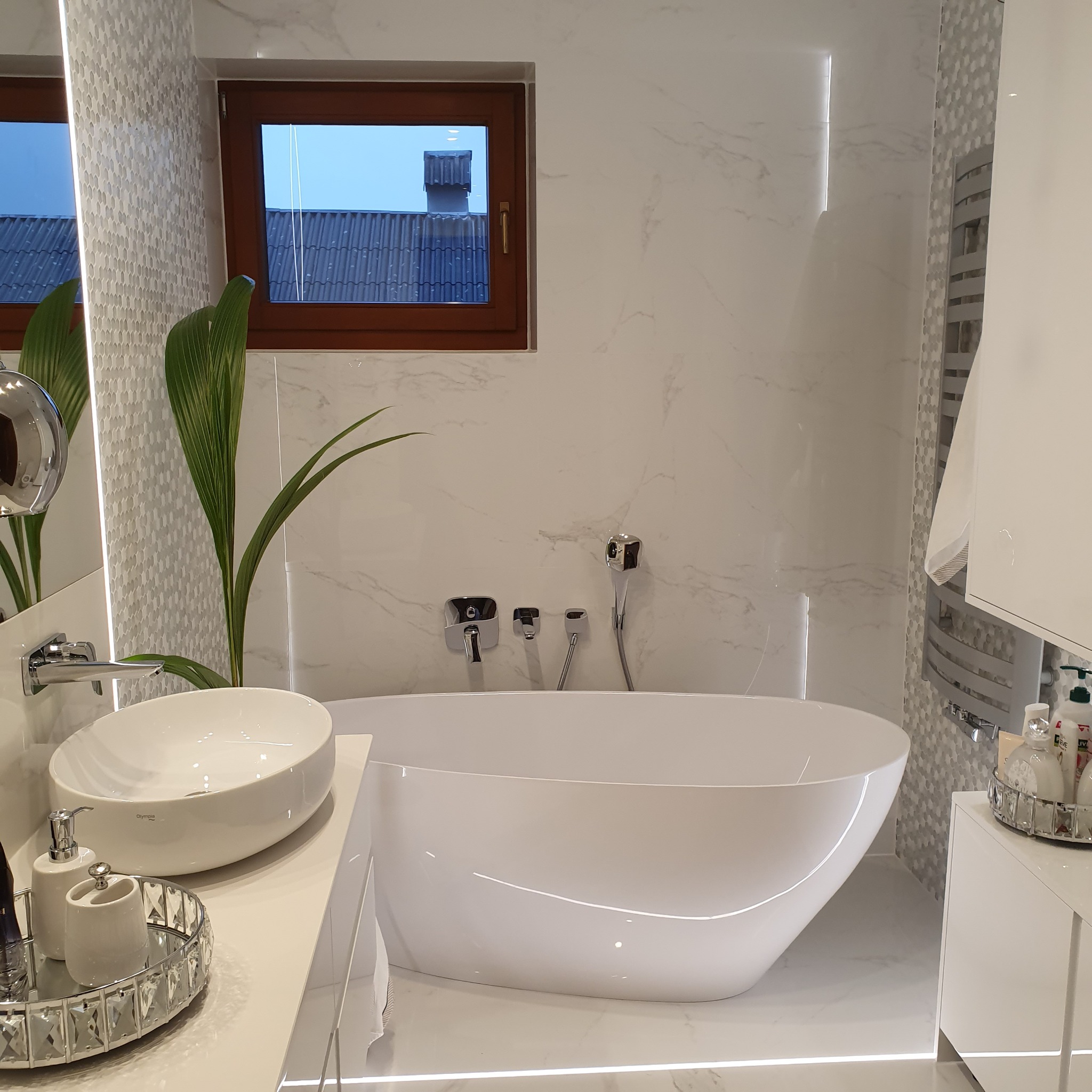 Nowoczesna łazienka w bieli z wolnostojącą wanną, umywalką nablatową i drewnianym oknem. W tle widoczny budynek.