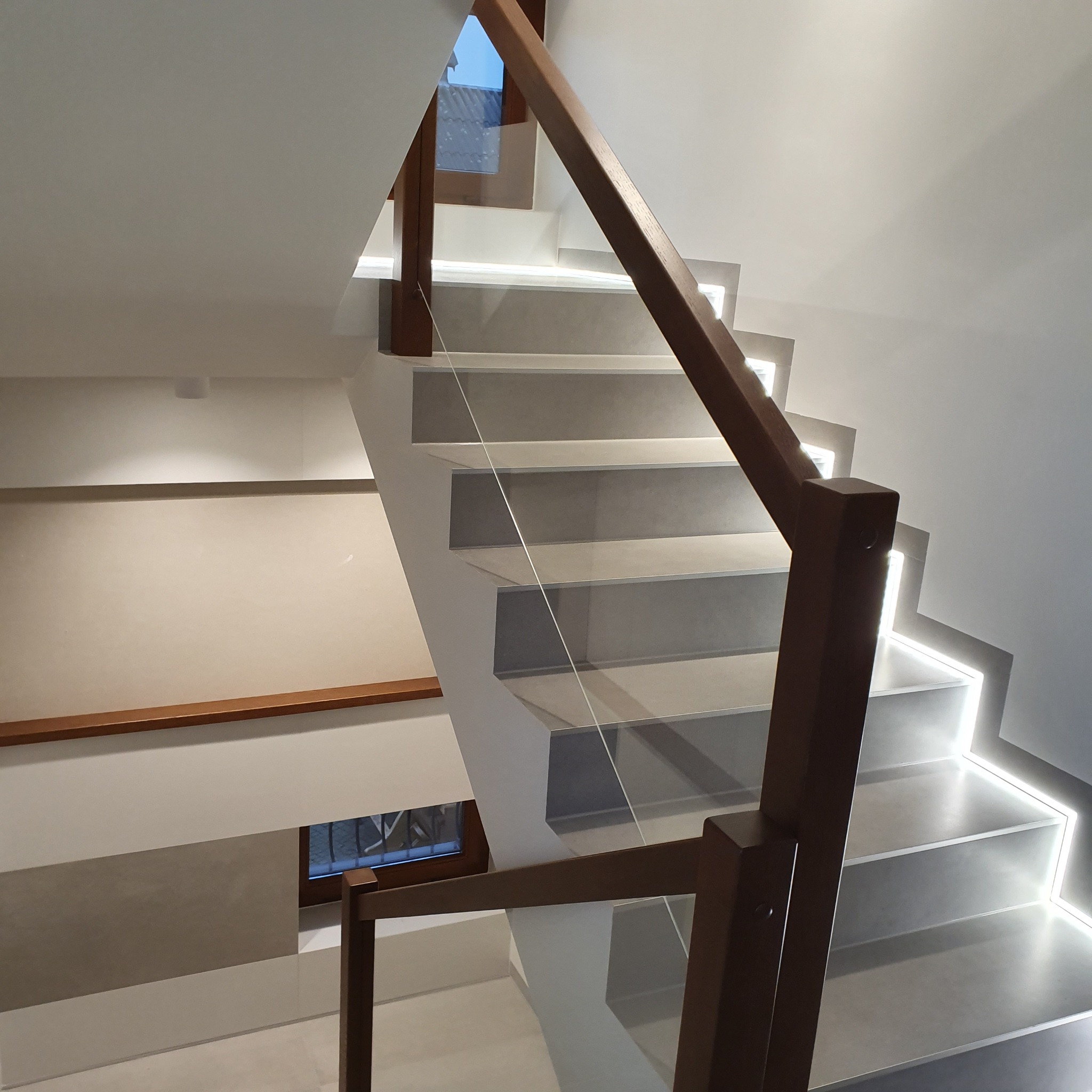 Nowoczesne schody z podświetleniem LED, drewnianą balustradą i szklanymi panelami. Widok z góry ukazuje minimalistyczny design i precyzyjne wykończenie.