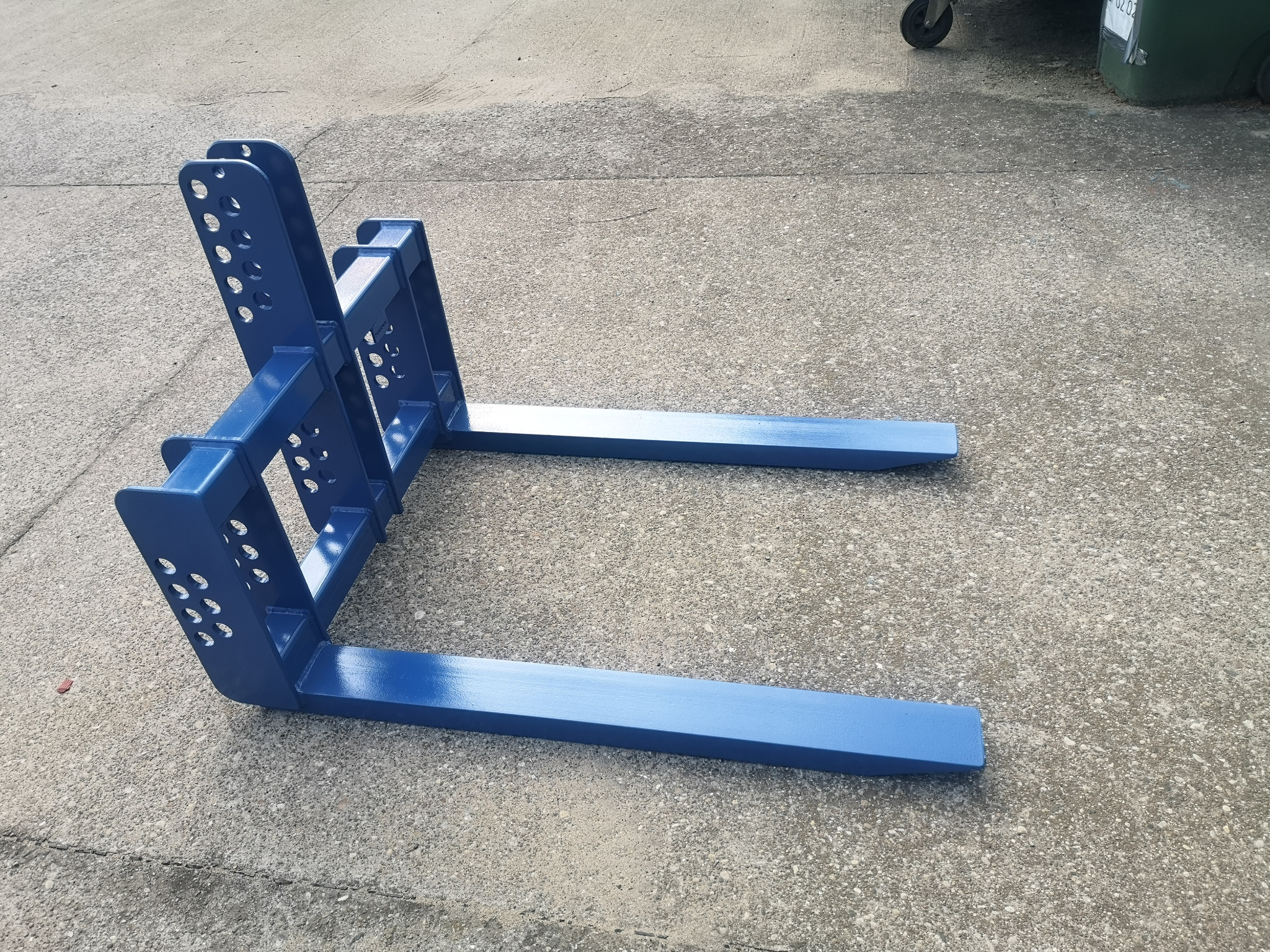 Niebieskie, metalowe widły do wózka widłowego, leżące na betonowym podłożu. Widoczne otwory w konstrukcji i ślady spawania.