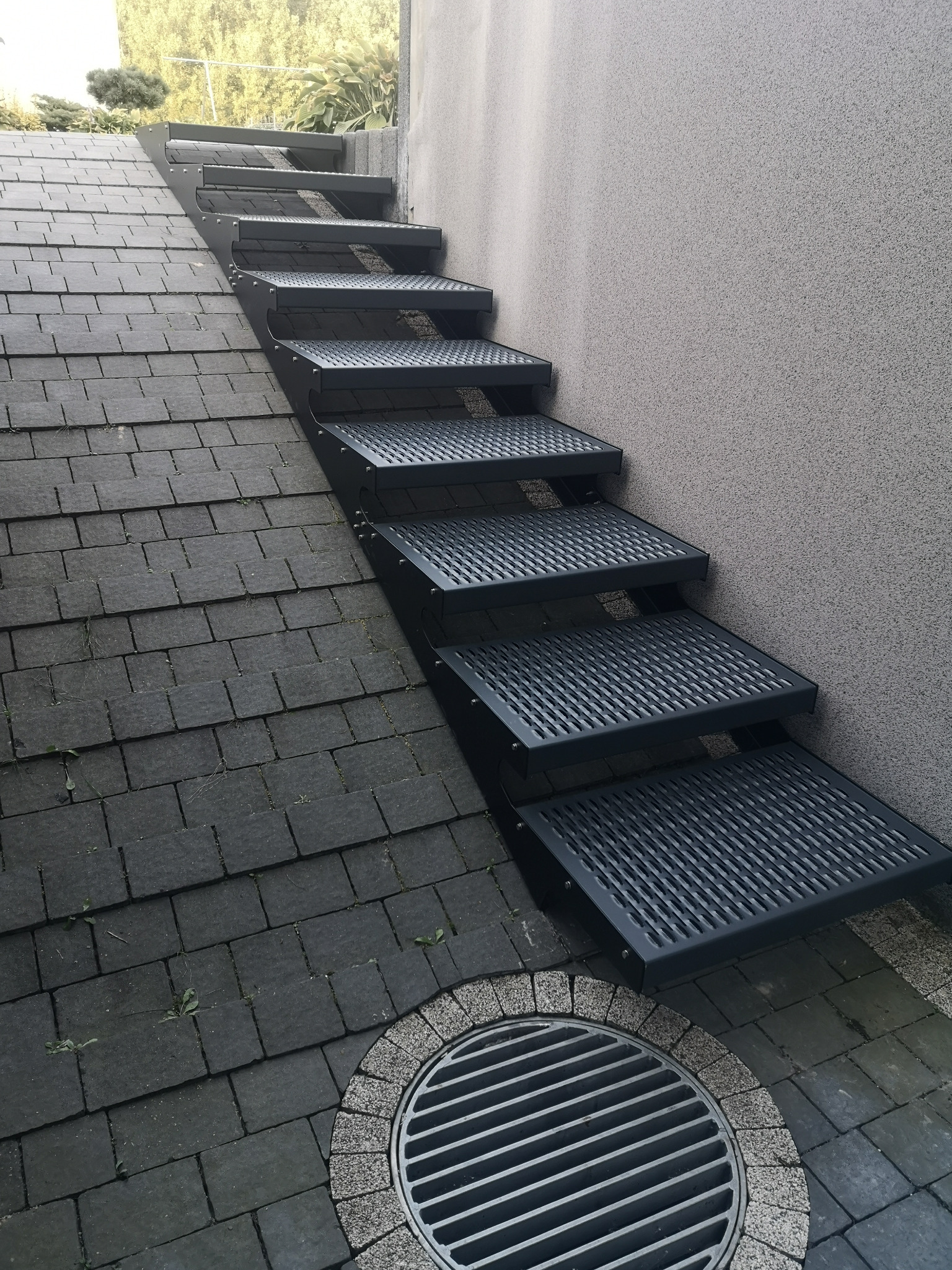 Nowoczesne, metalowe schody zewnętrzne z ażurowymi stopniami, prowadzące po nierównym terenie, przylegające do ściany budynku. Widoczny odpływ liniowy.