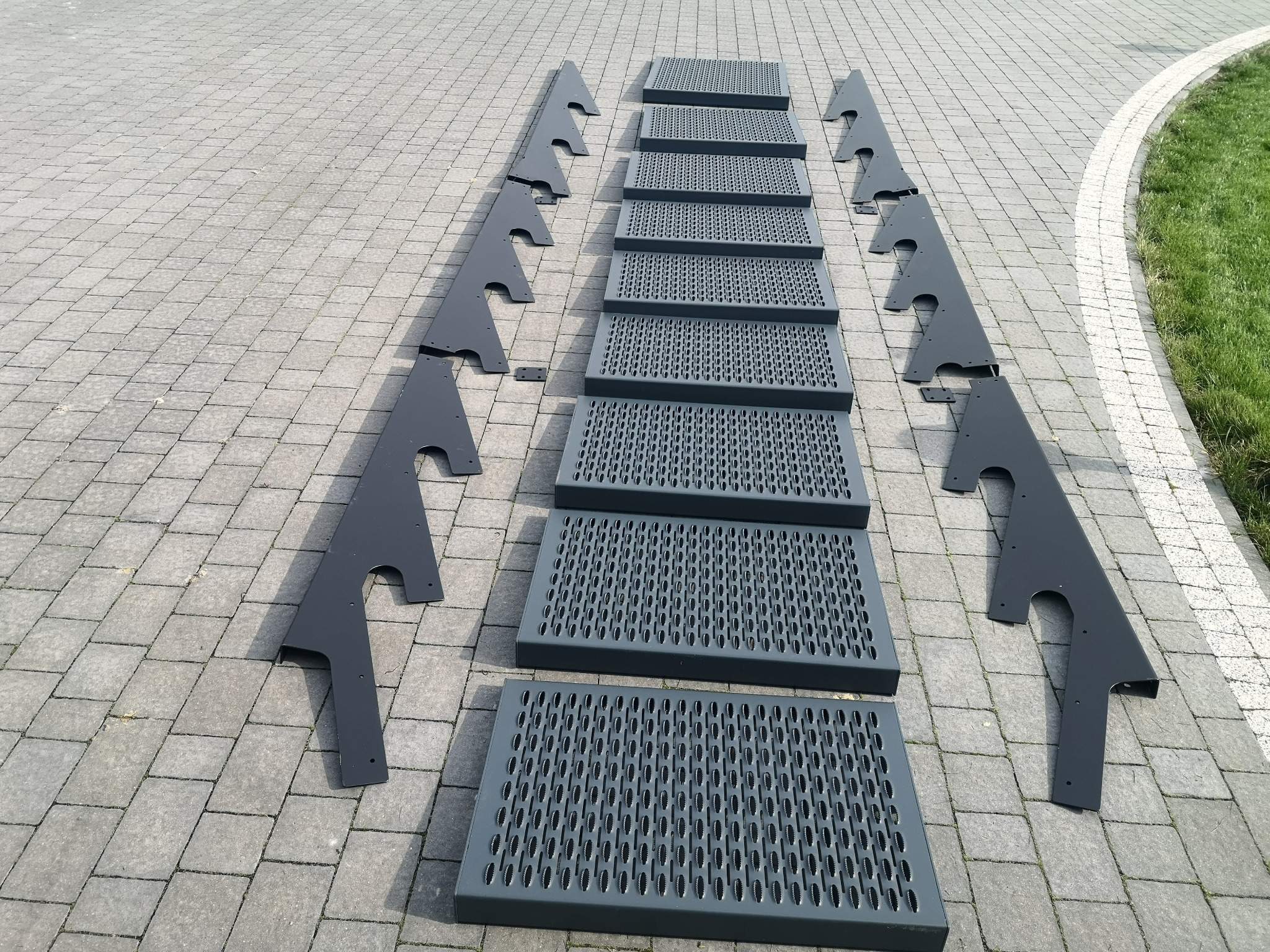 Metalowe stopnie schodowe i wsporniki w kolorze szarym, ułożone na brukowanej nawierzchni, gotowe do montażu. Widoczne perforacje na stopniach.