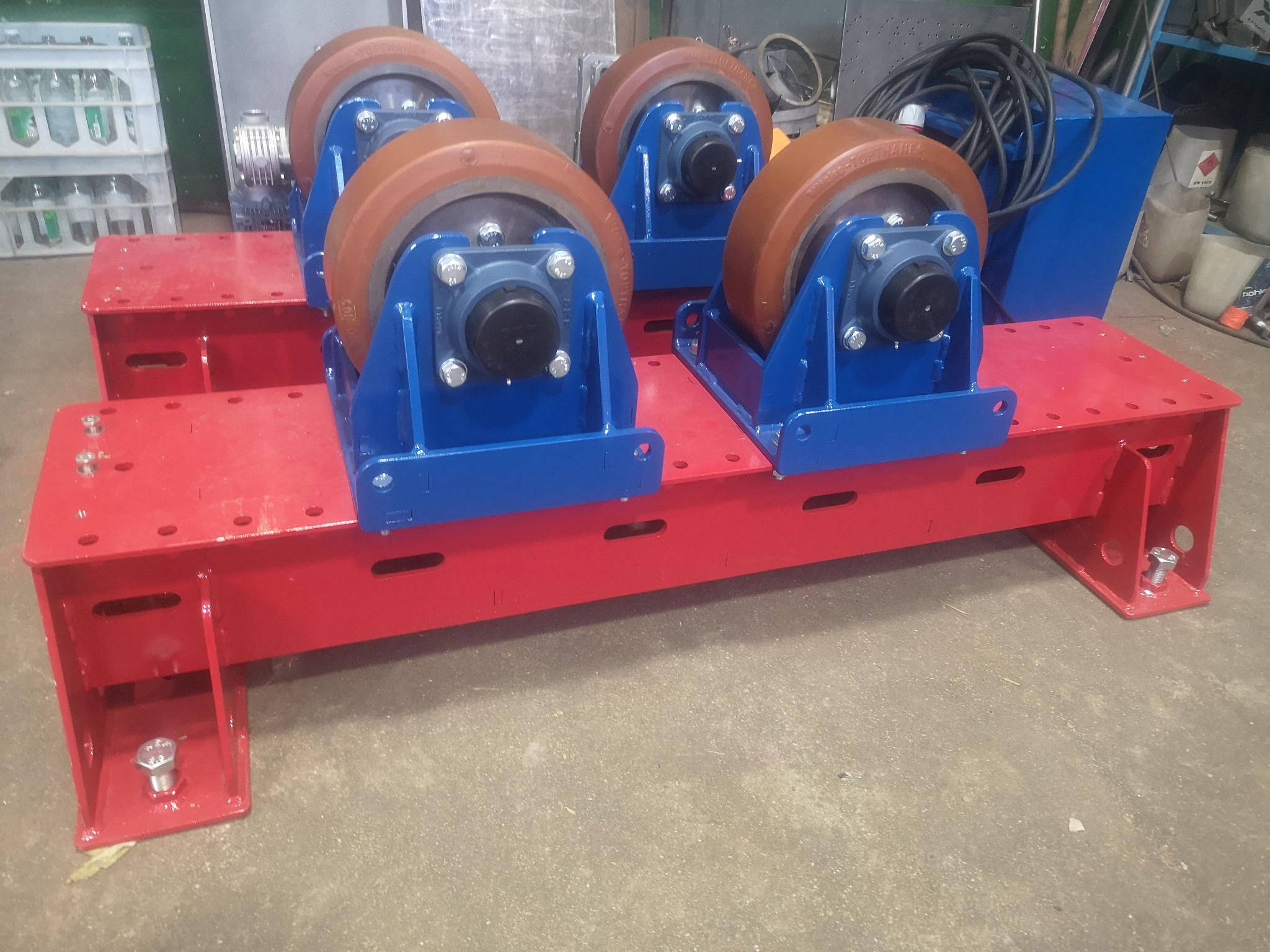 Czerwono-niebieski stojak rolkowy z czterema rolkami z brązowego poliuretanu, przeznaczony do obracania rur podczas spawania. Widoczne ślady spawania.