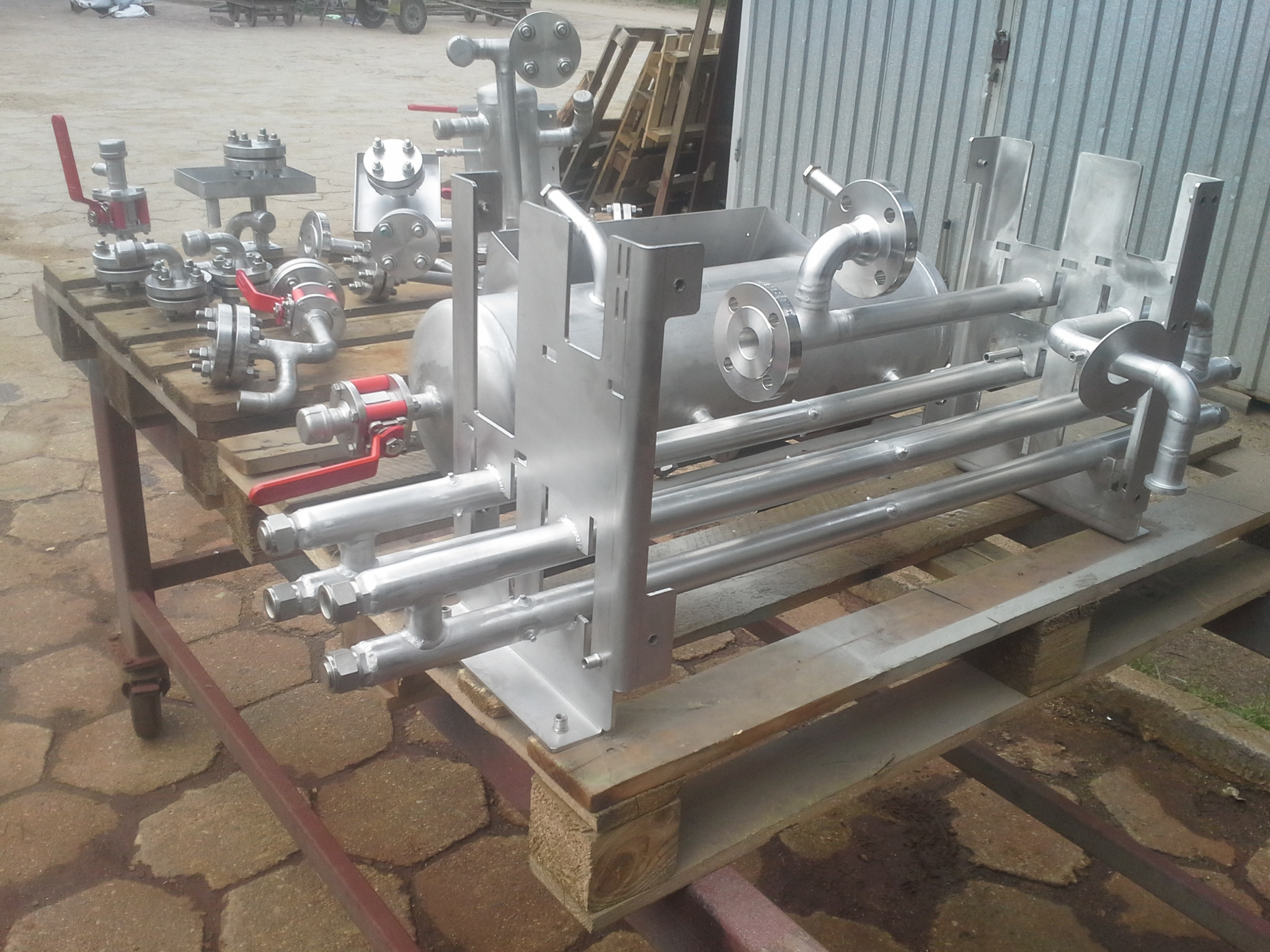 Srebrna, spawana konstrukcja z rur i zbiornika, umieszczona na drewnianych paletach. Elementy z czerwonymi zaworami z boku. Tło: blaszany garaż i elementy drewniane.