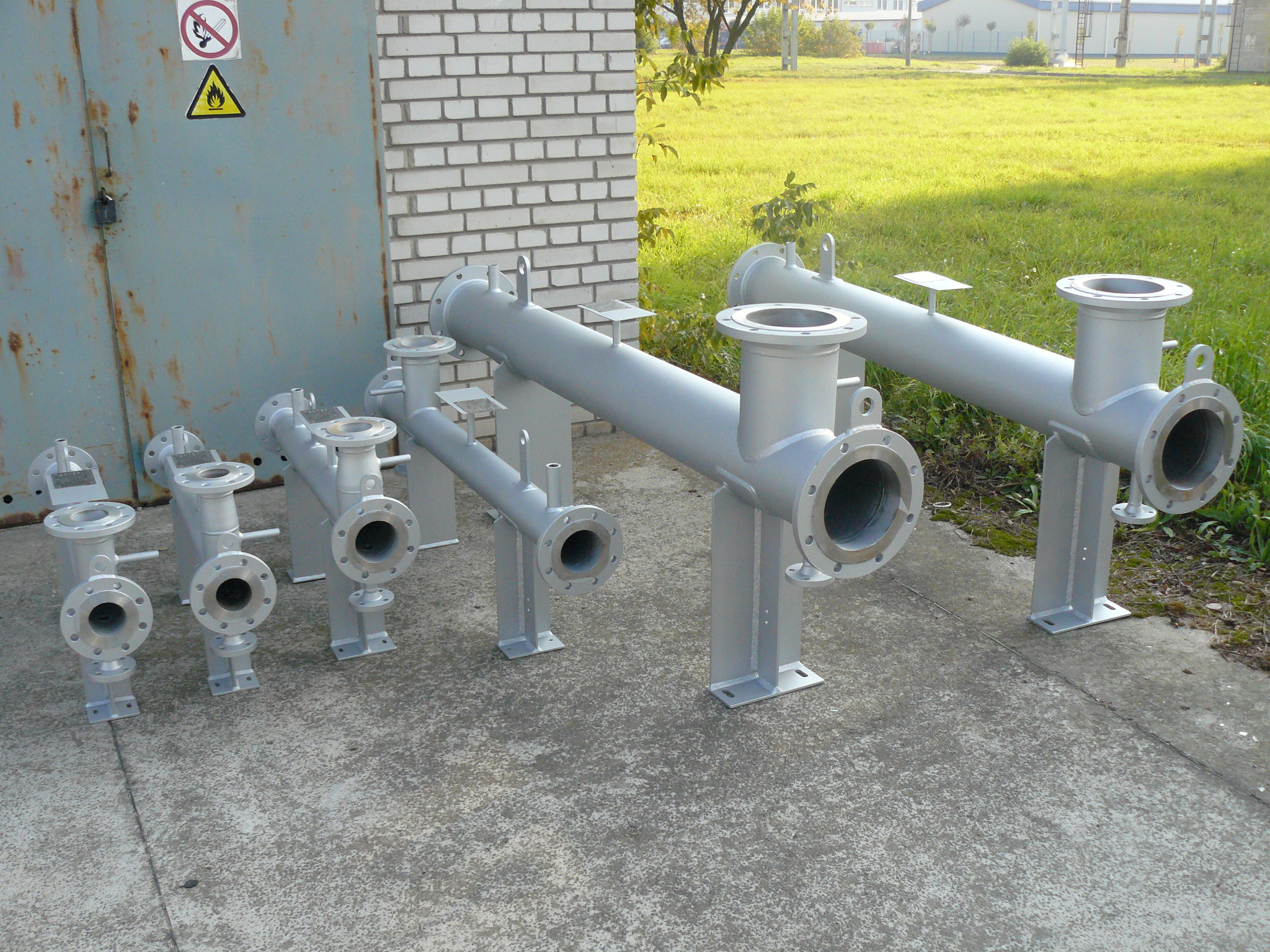 Szare, spawane rury z kołnierzami ustawione na betonowej powierzchni, z budynkiem w tle i znakiem ostrzegawczym na drzwiach.