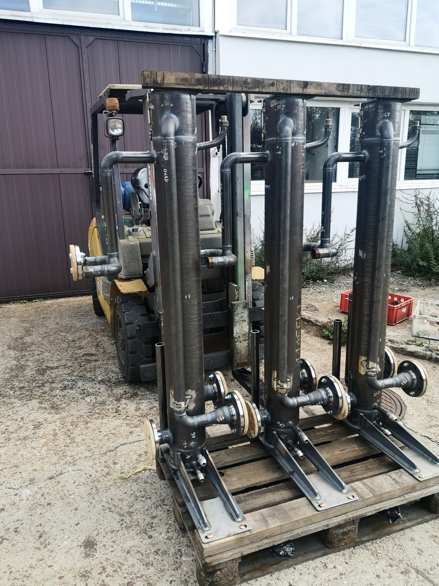 Trzy spawane, pionowe rury z przyłączami kołnierzowymi, umieszczone na palecie, w tle wózek widłowy i budynek z oknami. Widoczne spawy i oznaczenia na rurach.