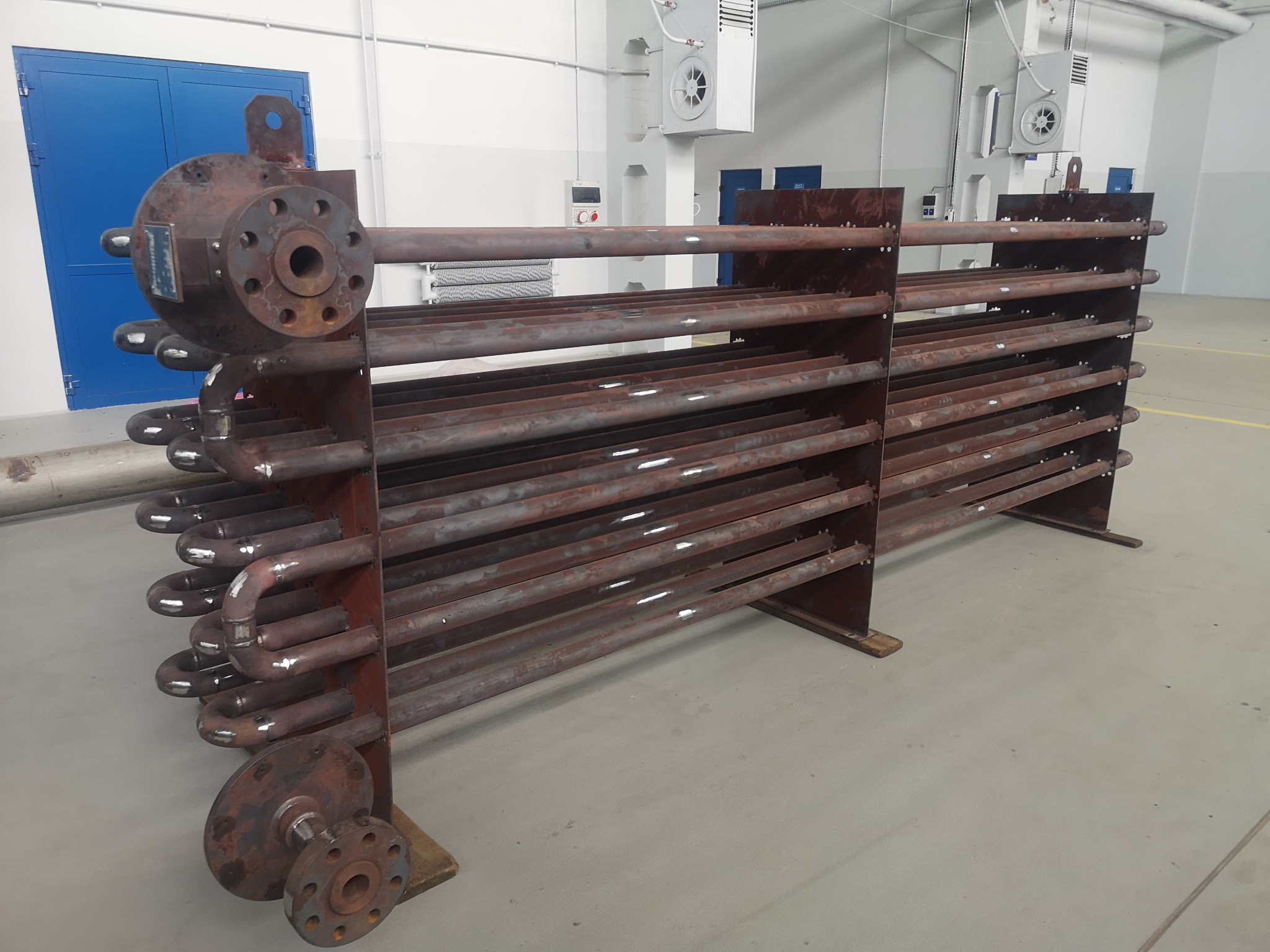 Spawana konstrukcja rurowa z kołnierzami, widoczne ślady po spawaniu. Konstrukcja metalowa w hali produkcyjnej, na szarym tle posadzki. Elementy wentylacji na ścianie.