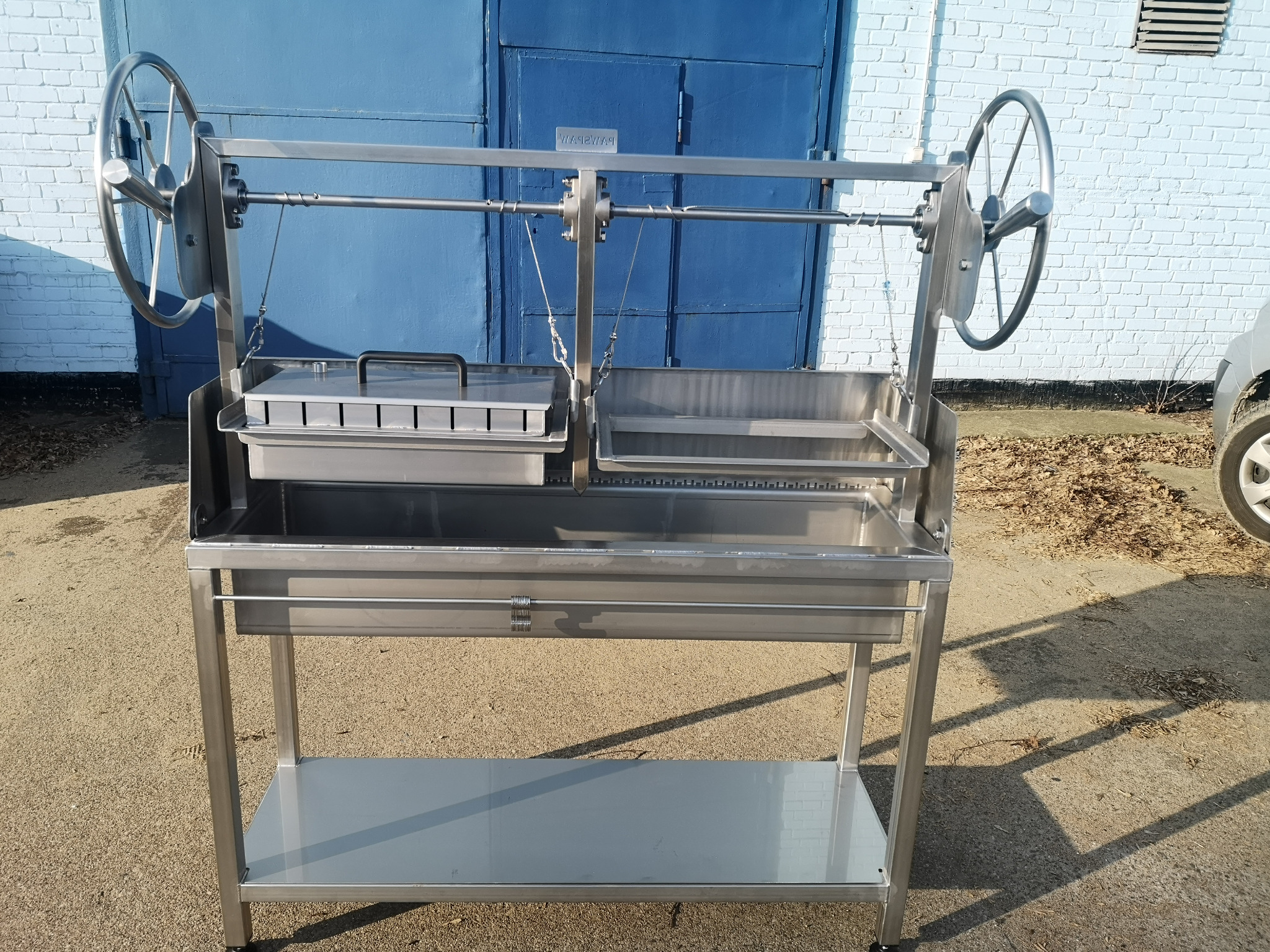 Metalowy grill z kołami do regulacji wysokości rusztu, na tle niebieskiej bramy i jasnej ściany z cegieł. Widoczna konstrukcja ze stali nierdzewnej.