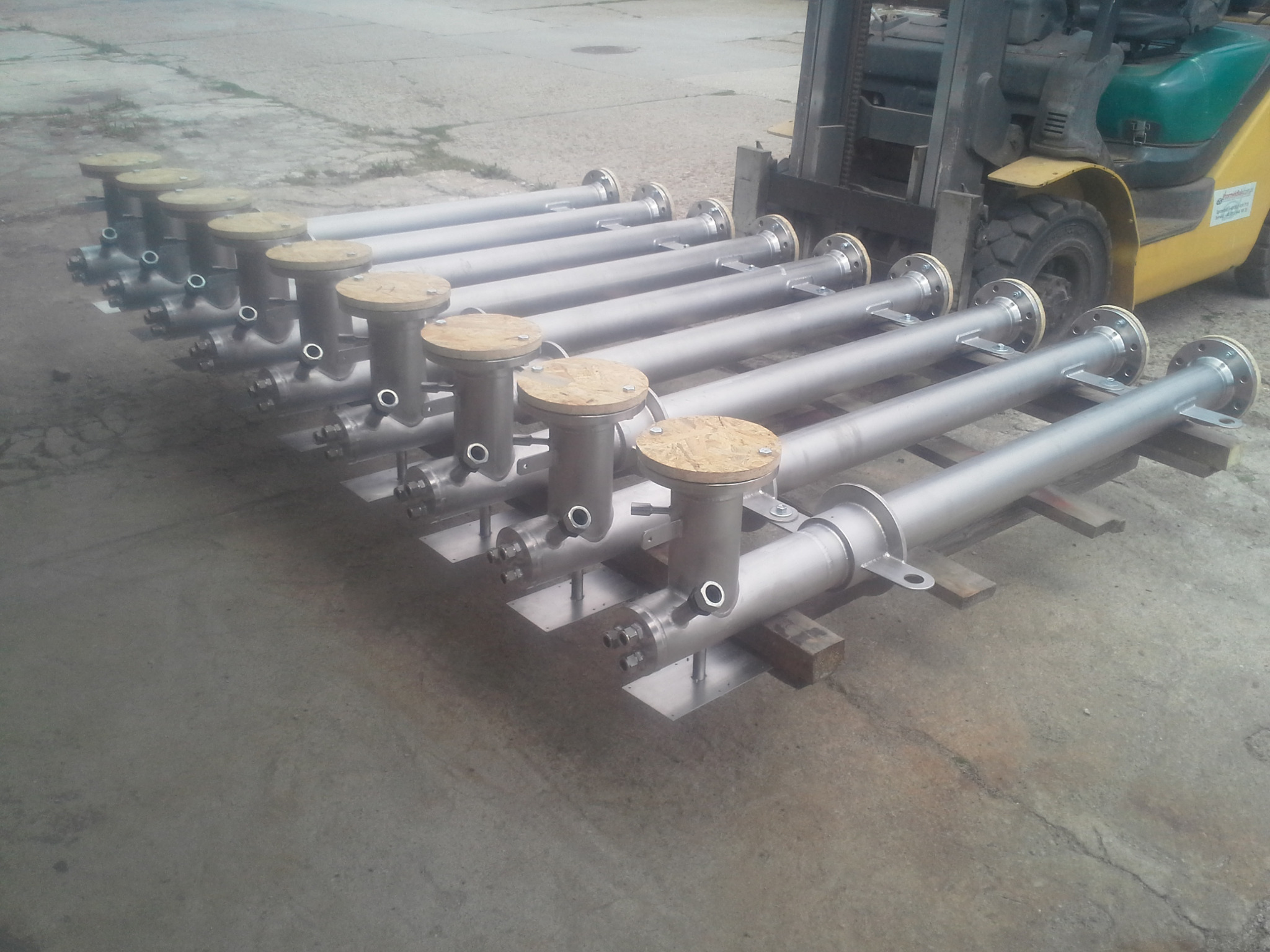 Rząd metalowych rur z kołnierzami i drewnianymi pokrywami, ułożonych na paletach, z żółtym wózkiem widłowym w tle. Precyzyjne spawy i wykończenie.