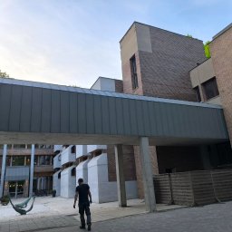 Siddach Spółka z o.o. - Szary, metalowy łącznik dachowy między budynkami z cegły, widziany z perspektywy pieszego. W tle dziedziniec z hamakiem.