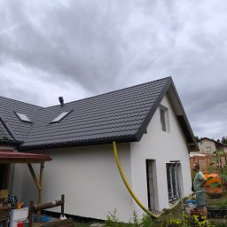 Siddach Spółka z o.o. - Dom z szarą dachówką i żółtą rurą spustową. Widoczny fragment elewacji, okna w trakcie montażu i osoba stojąca obok pomarańczowej betoniarki. Zachmurzone niebo w tle.