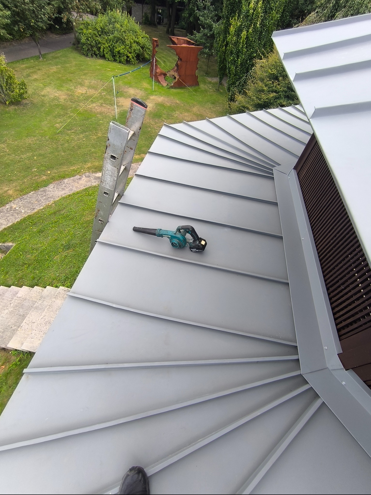 Szary, metalowy dach z widoczną dmuchawą akumulatorową Makita, drabina oparta o dach, widok z góry na posesję z zielenią i rzeźbą.