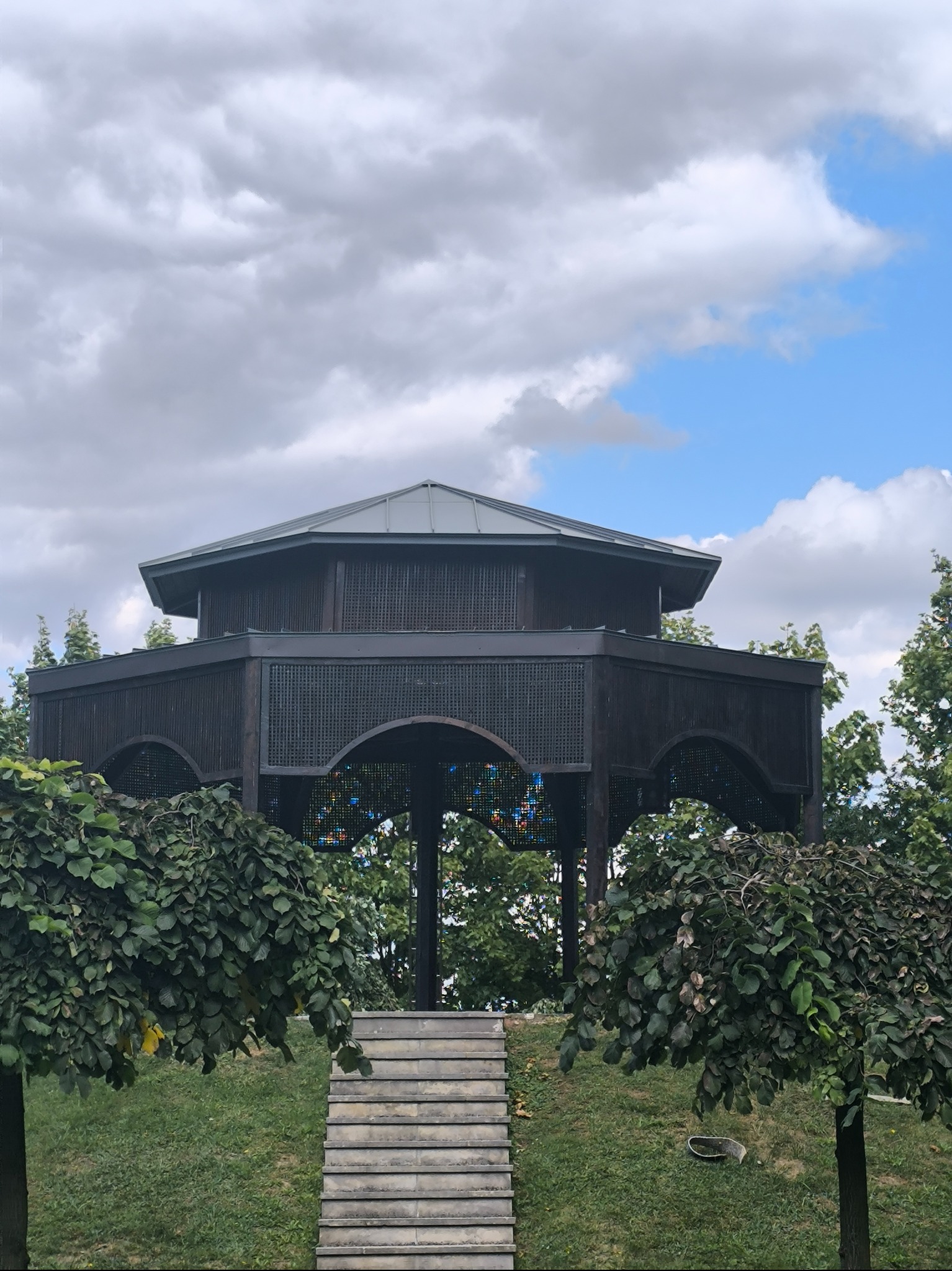 Ośmioboczna altana z ciemnego drewna i szarym dachem, widziana z dołu, z kamiennymi schodami i ozdobnymi drzewami w tle, pod pochmurnym niebem.