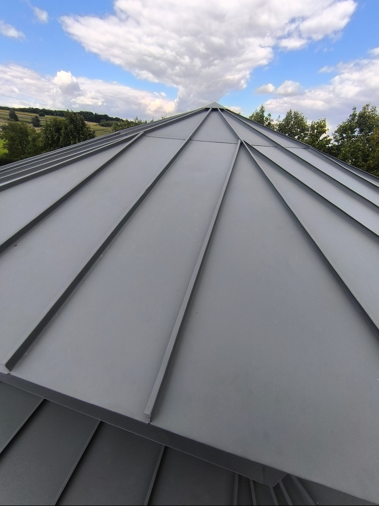 Szary dach z blachy trapezowej w zbliżeniu, widok z dołu, na tle błękitnego nieba i zieleni. Widoczne symetryczne ułożenie blach i estetyczne wykończenie.