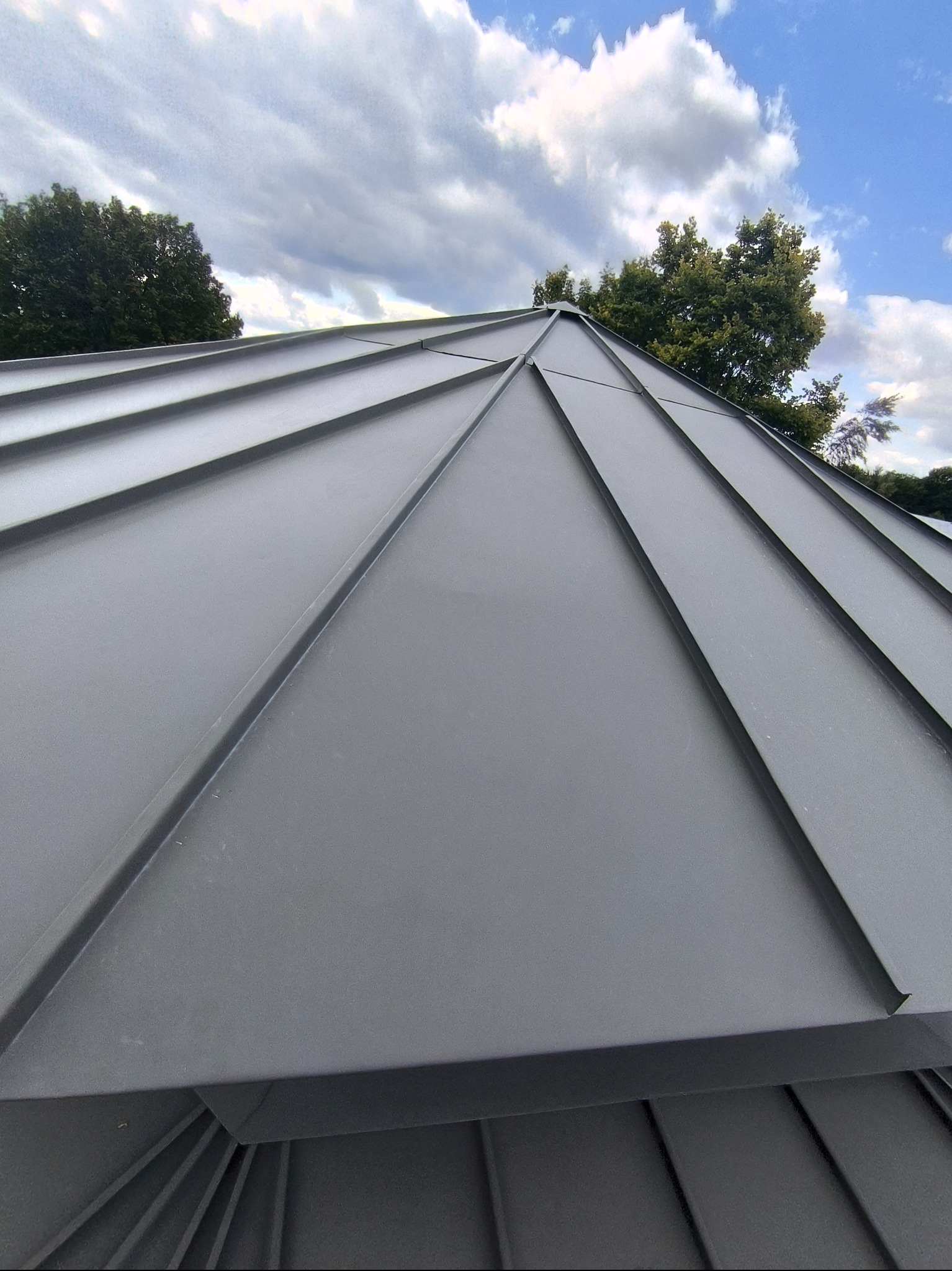 Szary, metalowy dach z wyraźnymi liniami łączeń blach, widziany pod kątem z dołu, na tle błękitnego nieba z chmurami i zieleni drzew.