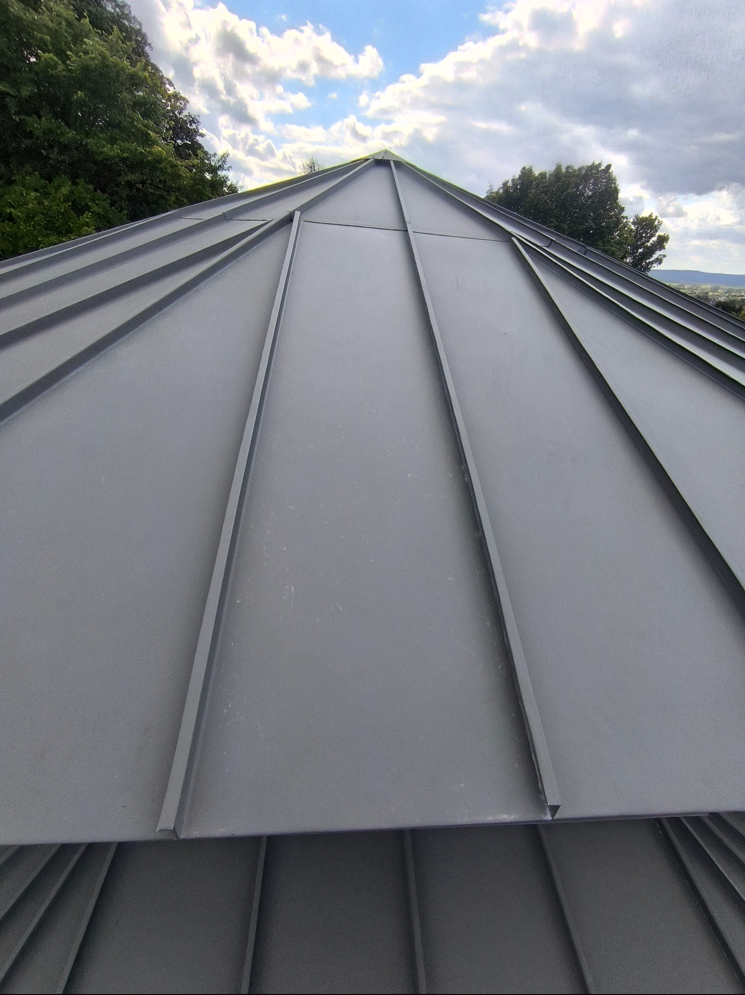 Szary dach z blachy trapezowej z widocznymi łączeniami na tle błękitnego nieba i zieleni drzew. Precyzyjne wykonanie, nowoczesny design.