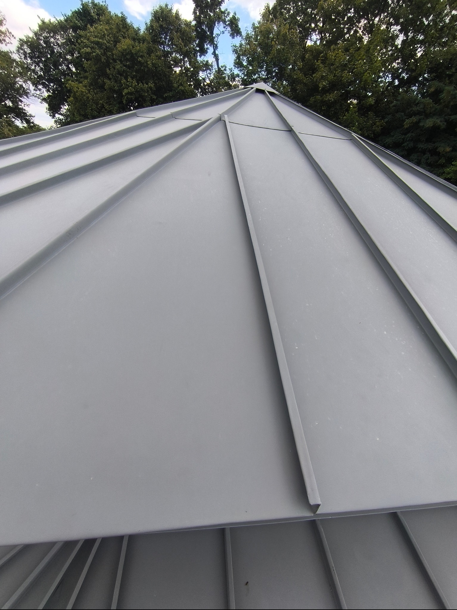 Szary dach z blachy trapezowej, widok z dołu, z zielenią drzew w tle. Precyzyjne łączenia blacharskie, regularny układ paneli. Nowoczesny design.
