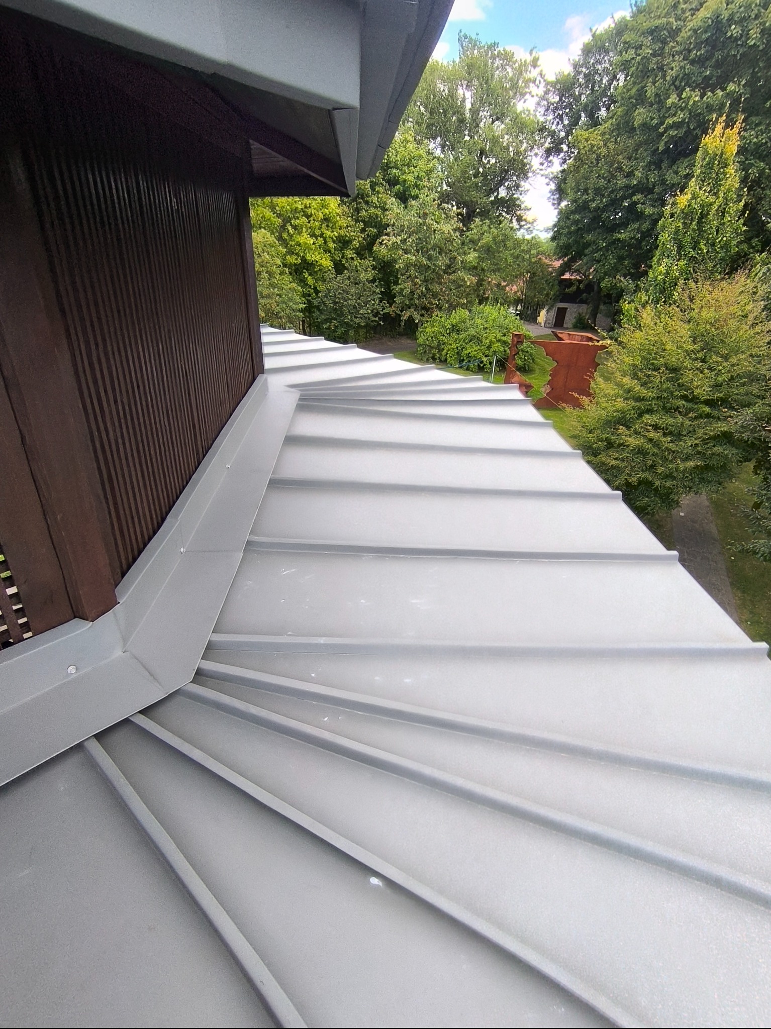 Szary dach z blachy na budynku z brązową elewacją, widok z góry, zieleń w tle. Detal obróbek blacharskich i łączenia dachu z elewacją. Nowoczesne krycie dachu.