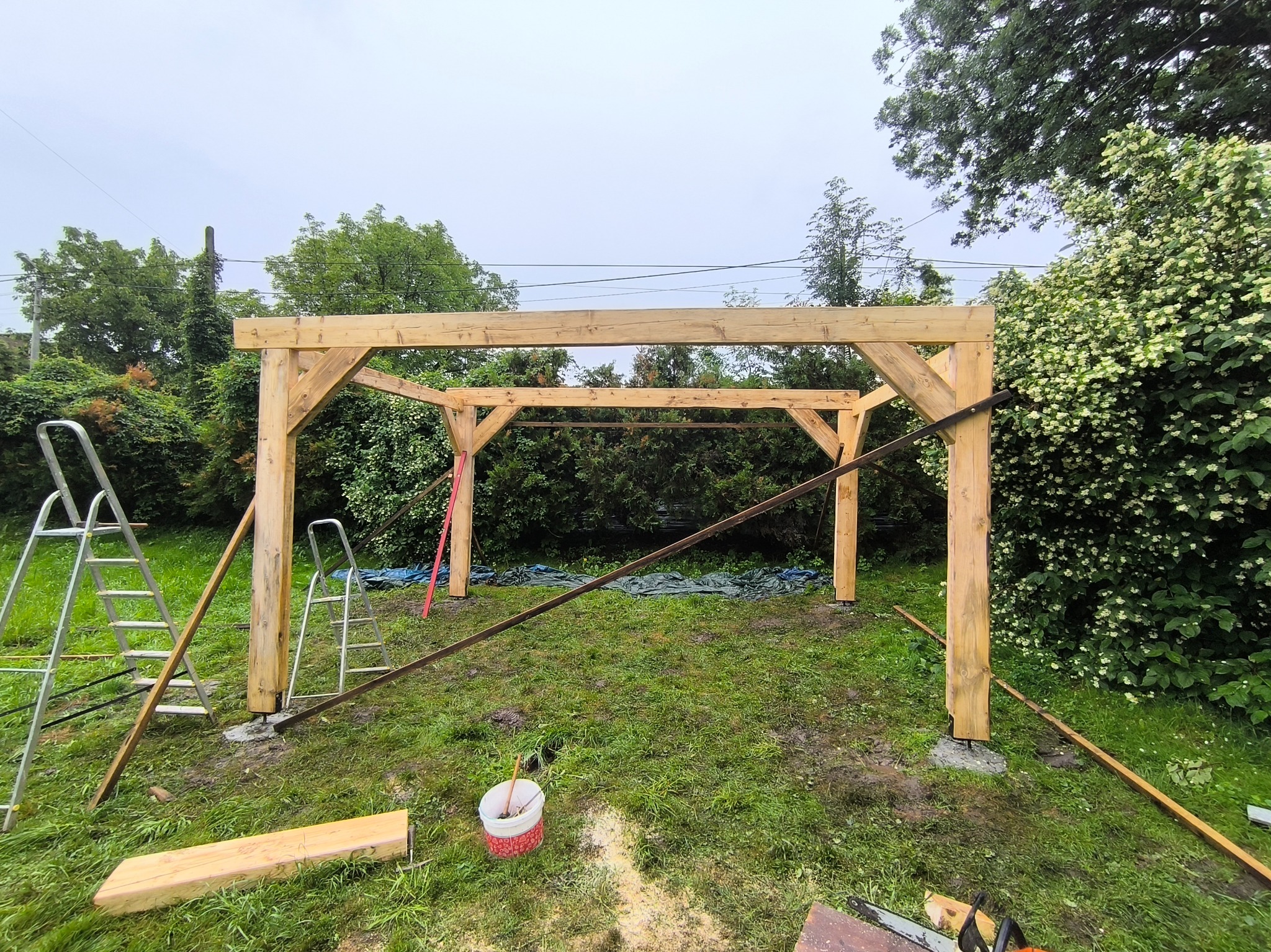 Drewniana konstrukcja dachu w trakcie budowy, widoczne rusztowania i narzędzia na trawiastym terenie. Etap montażu belek i słupów w ogrodzie.